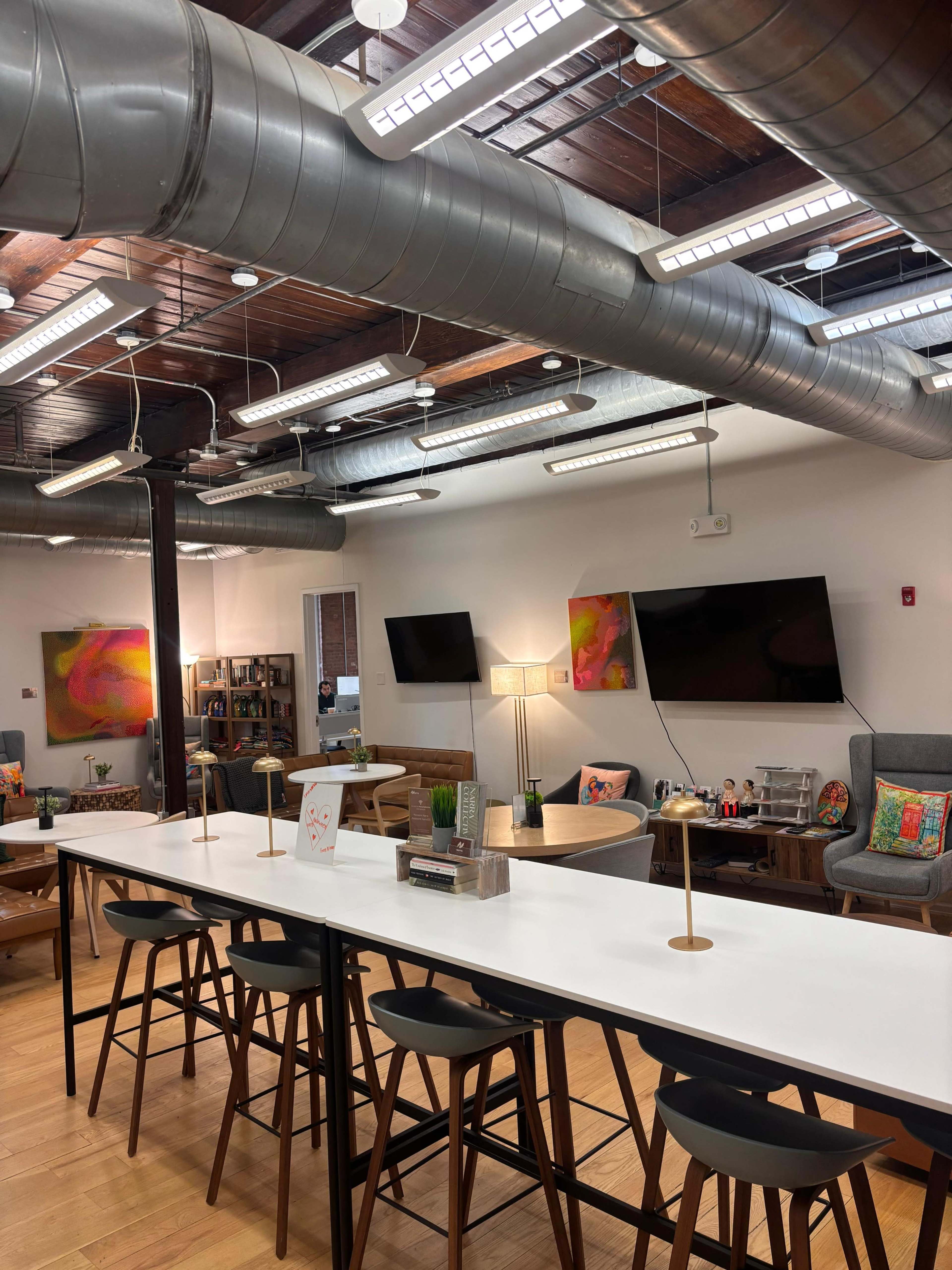 The image shows a modern workspace featuring a long white table with black bar stools, exposed ductwork on the ceiling, and colorful artwork on the walls.