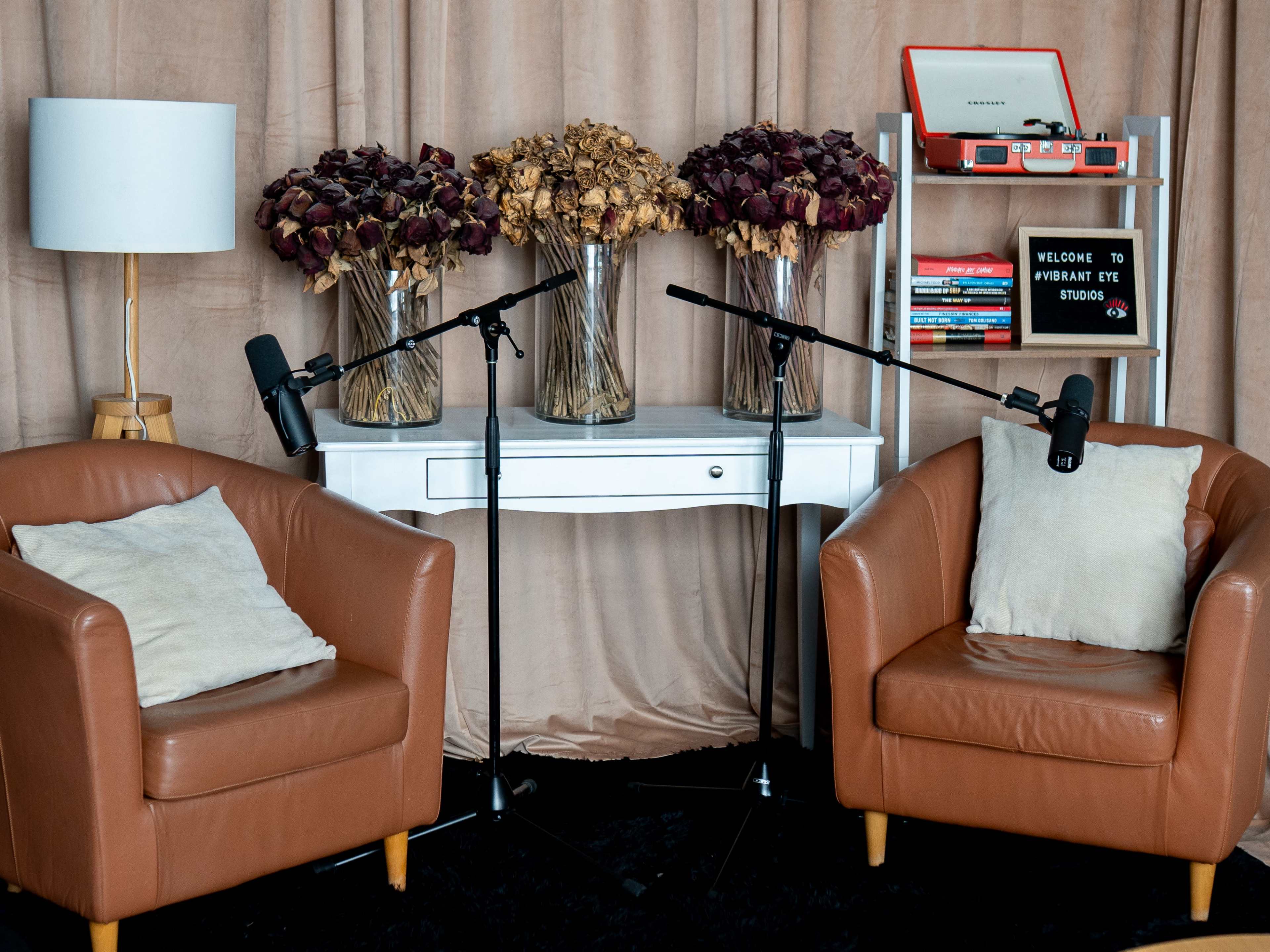 A cozy studio setup features two brown leather chairs, a white console table, two microphones on stands, and three vases of dried flowers against a backdrop of beige fabric.