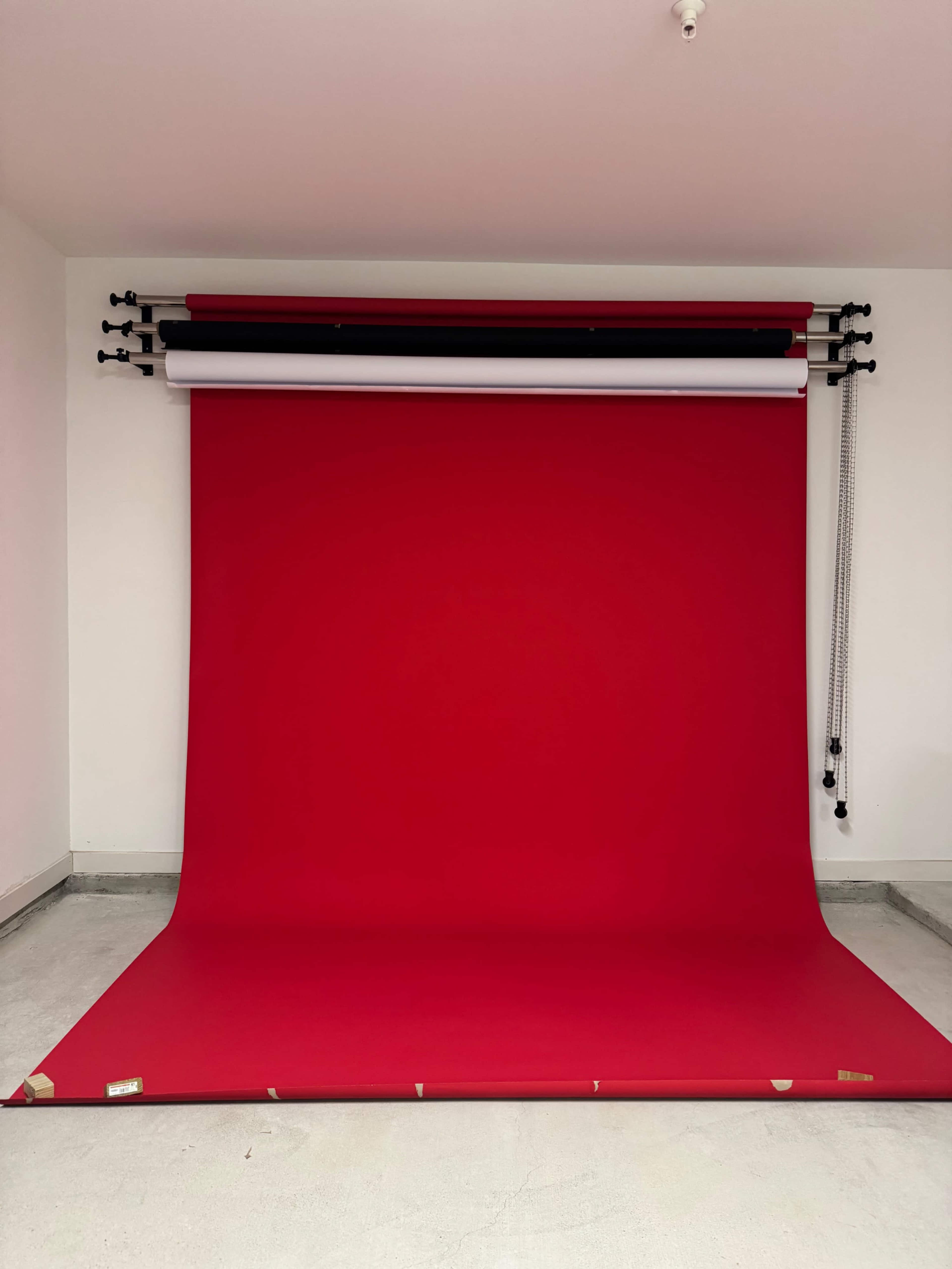 The image shows a photography backdrop setup featuring a large red fabric roll, with additional rolls of white and black hanging above it on a support system.
