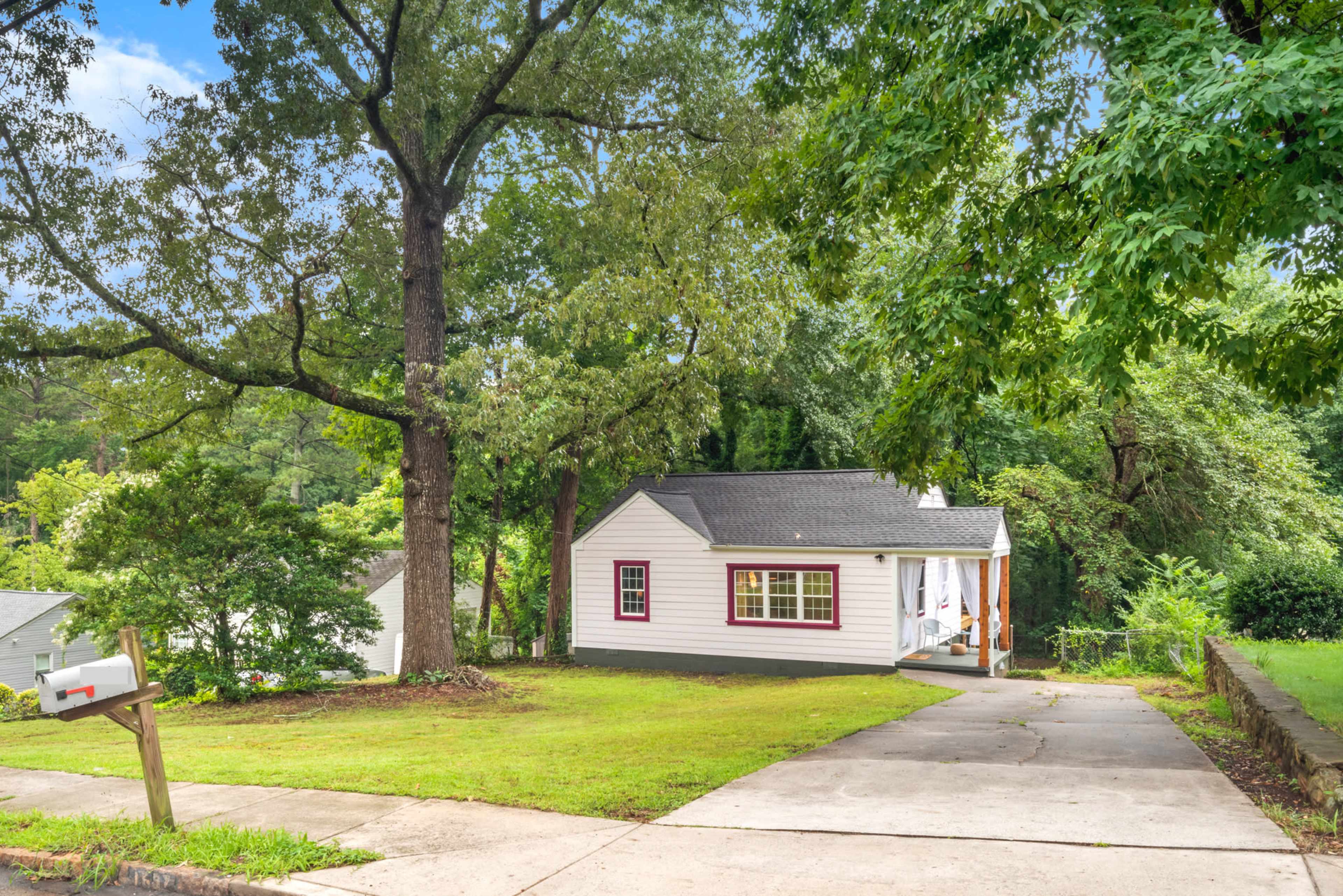 A small white house with maroon trim is situated on a sloped lawn, surrounded by greenery, with a driveway leading up to it.