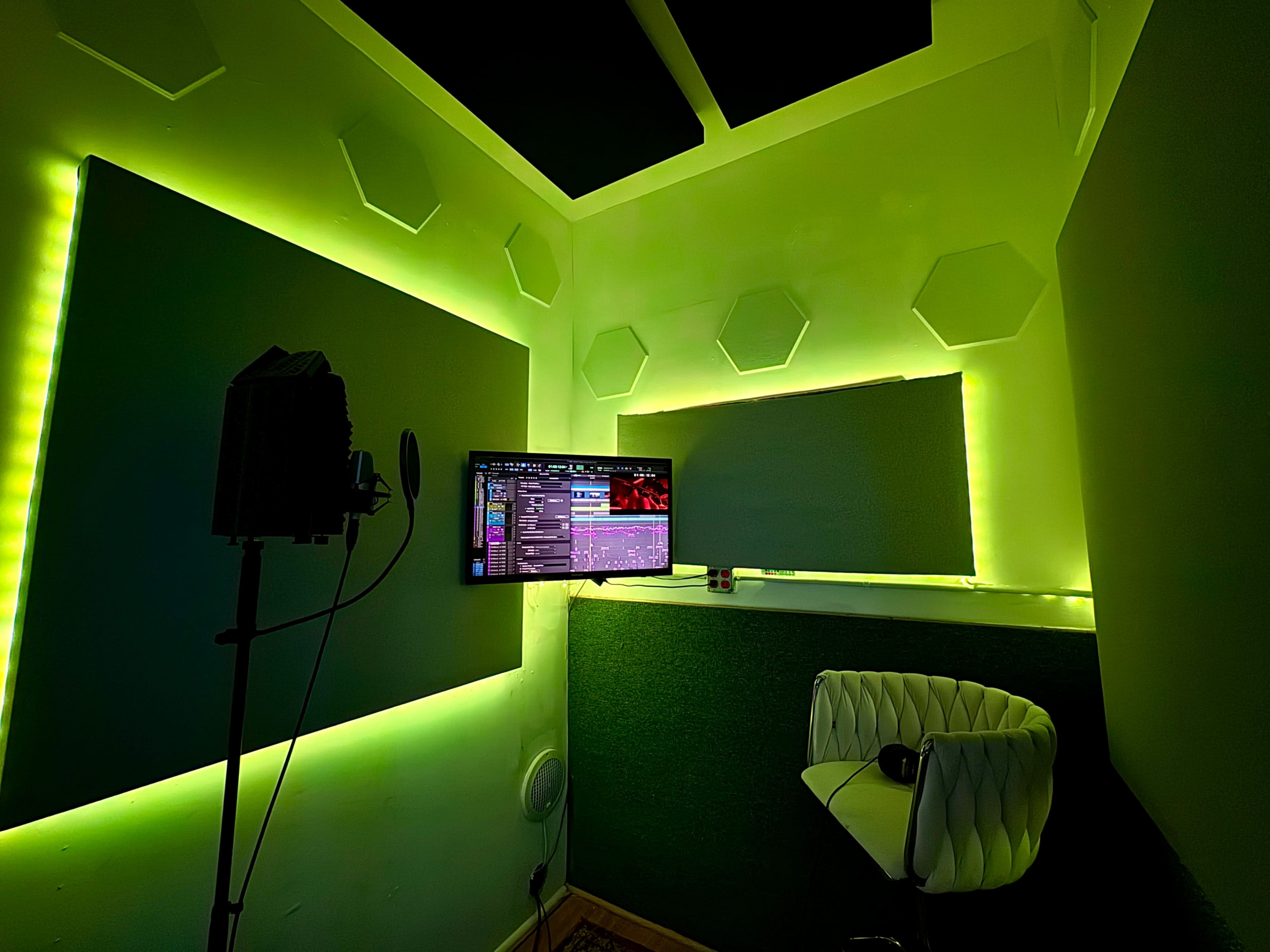 The image shows a small corner room equipped with a microphone on a stand, a wall-mounted monitor, and green LED lighting illuminating the space.