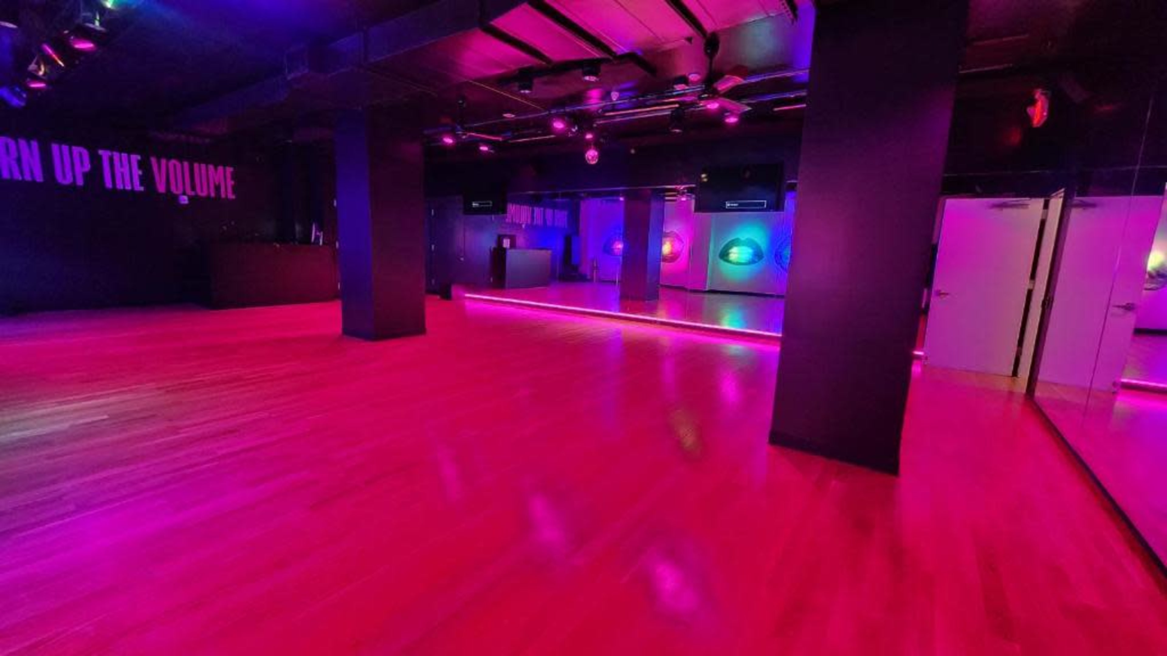 A spacious dance studio features polished wooden floors illuminated by purple and pink lights, with walls that include modern sound equipment.