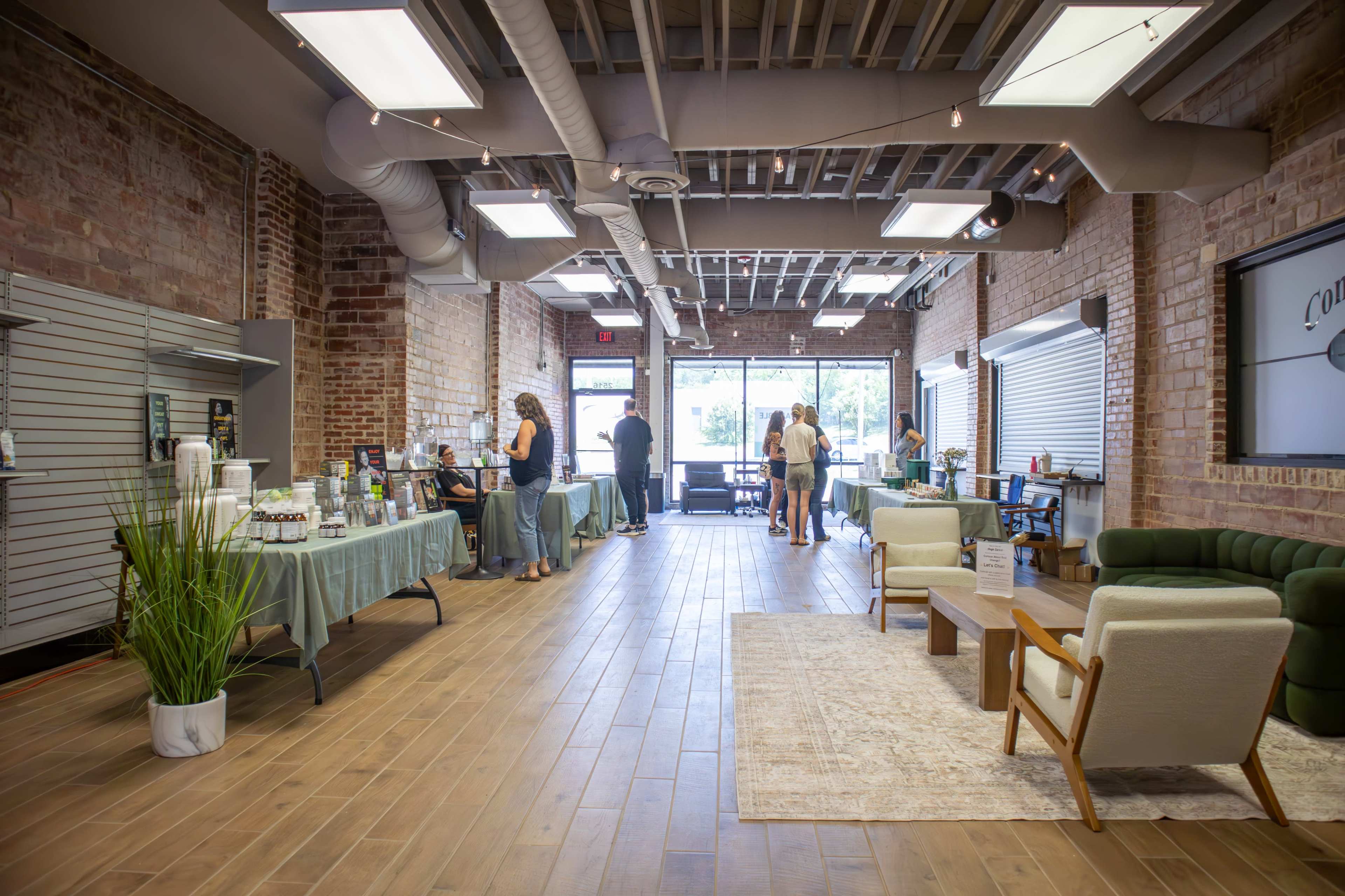 The image shows a spacious, modern retail space with exposed brick walls, wooden flooring, and various display tables set up for products, while people interact at the front of the shop.