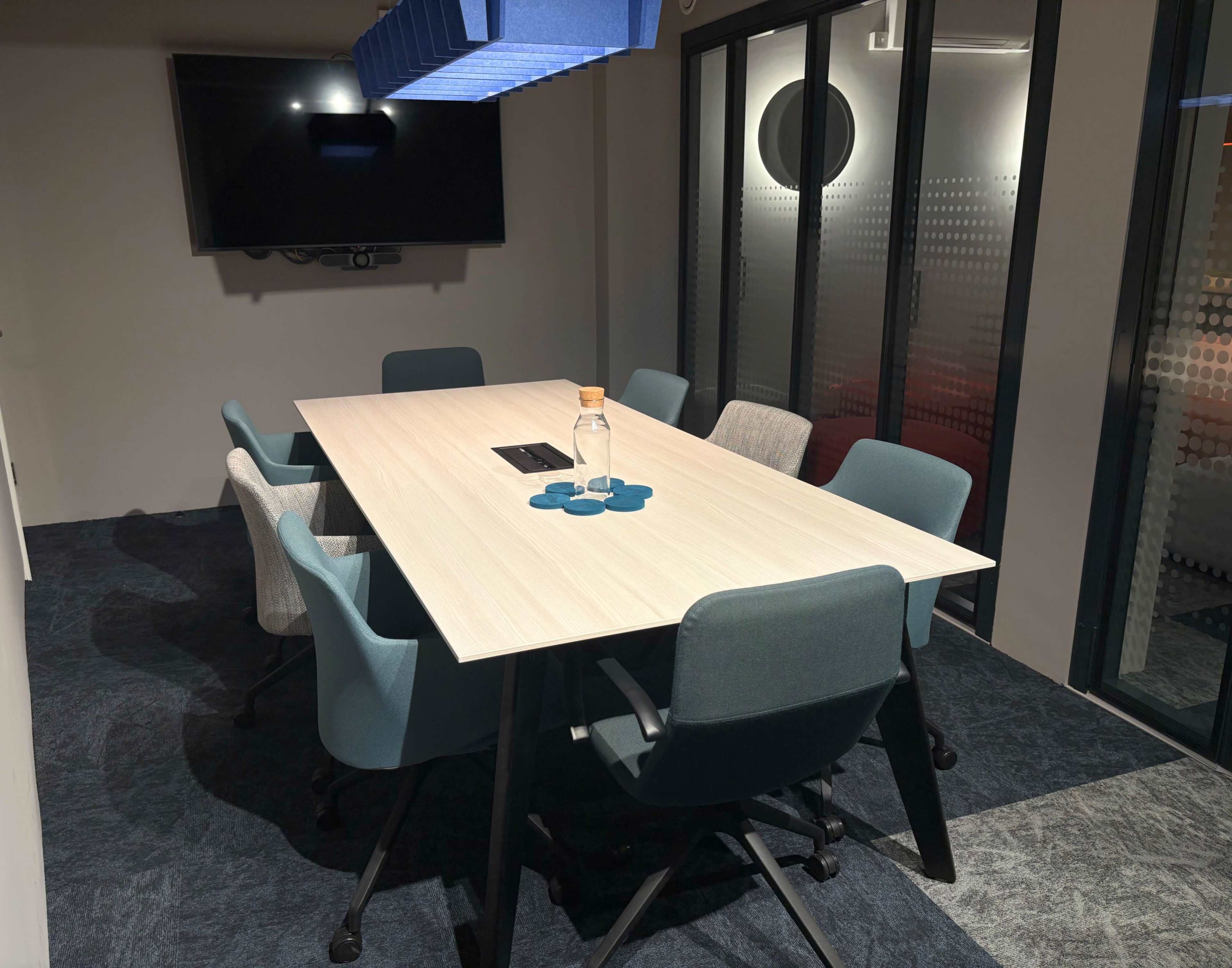 A modern conference room features a large table surrounded by several chairs, with a television mounted on the wall and glass doors.