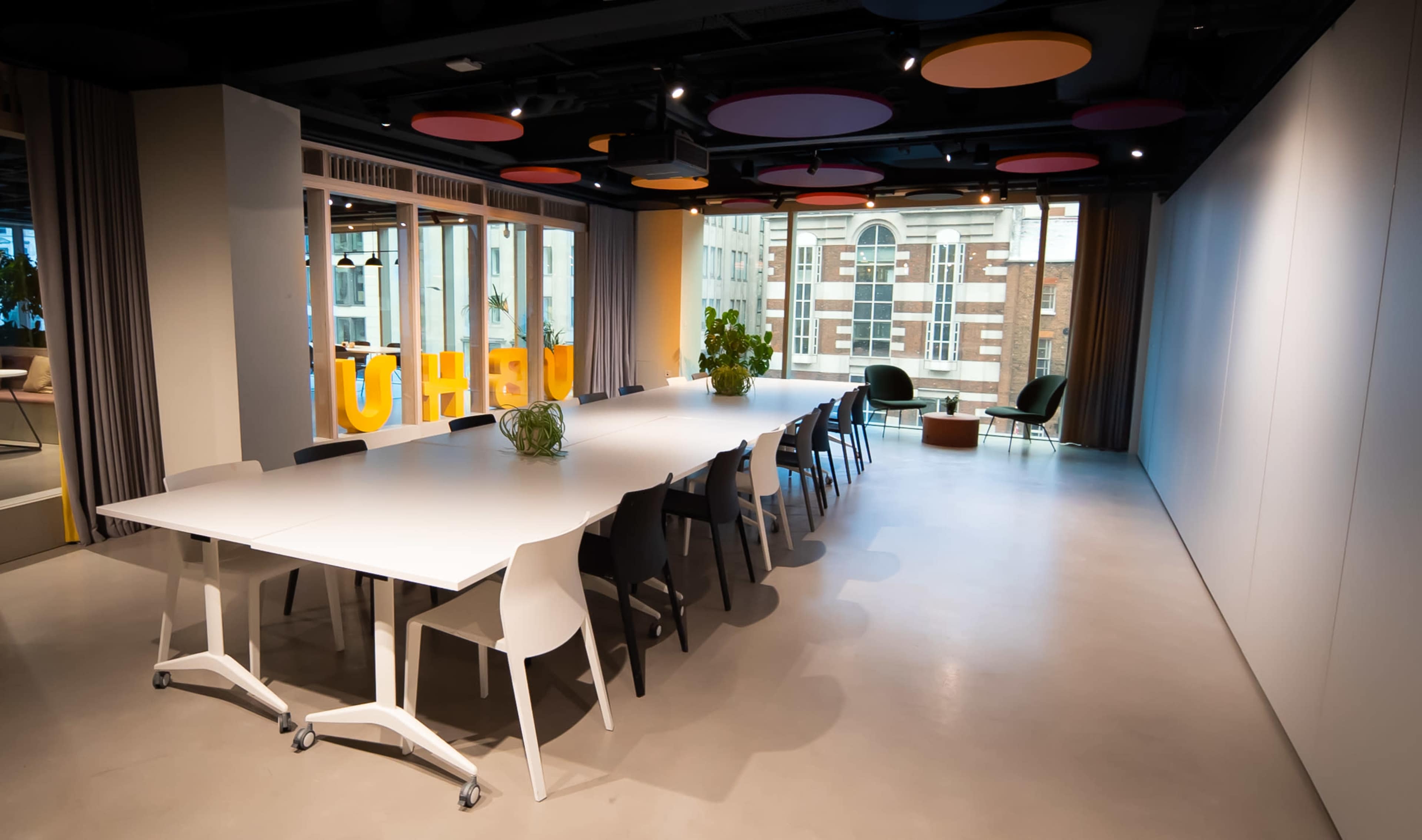 A modern conference room features a large white table surrounded by chairs, with colorful acoustic panels on the ceiling and a view of the city through large windows.