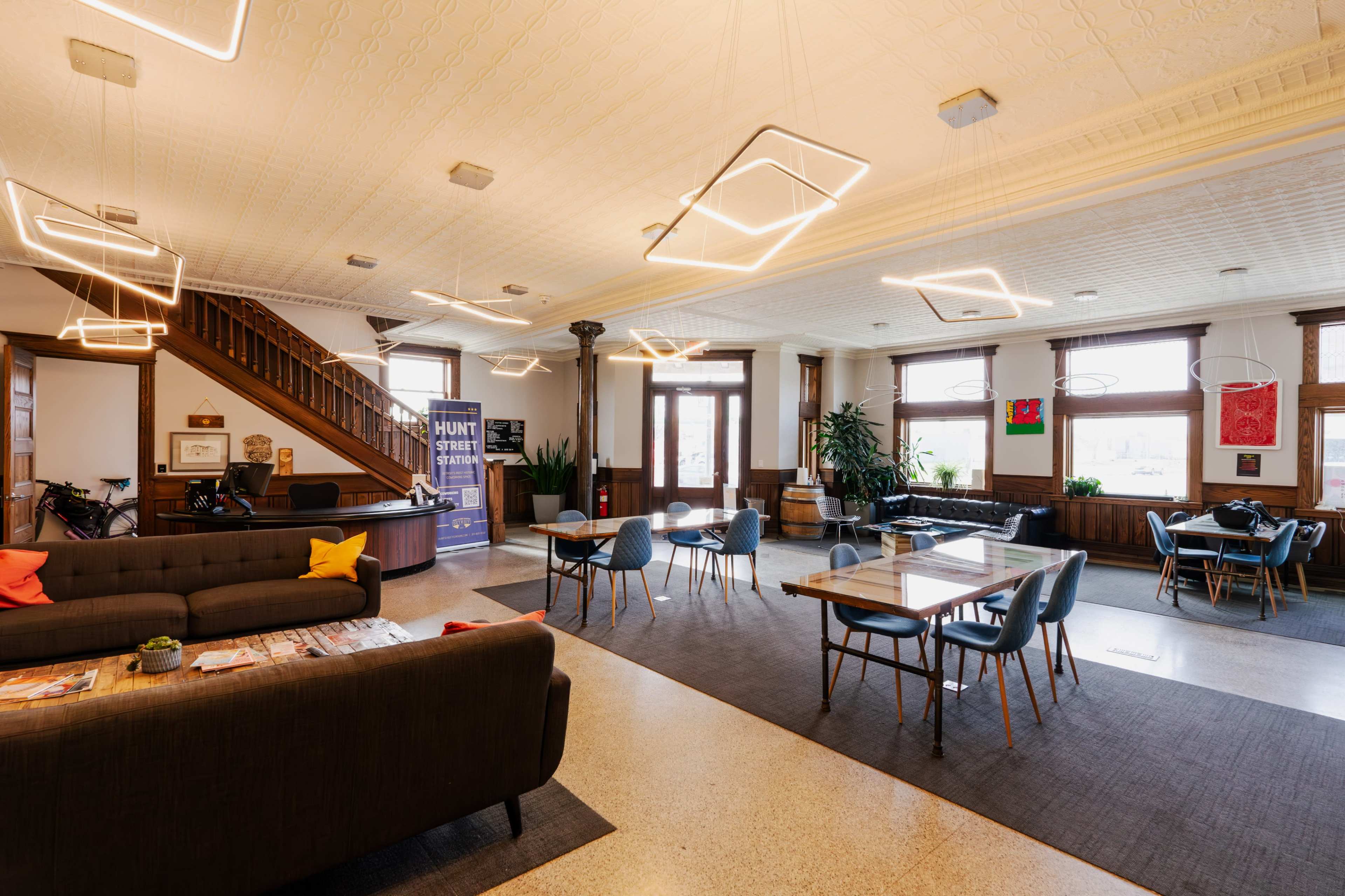A spacious lobby features modern furniture, including a dark sofa and light wooden tables, with unique lighting fixtures and large windows allowing natural light to enter.