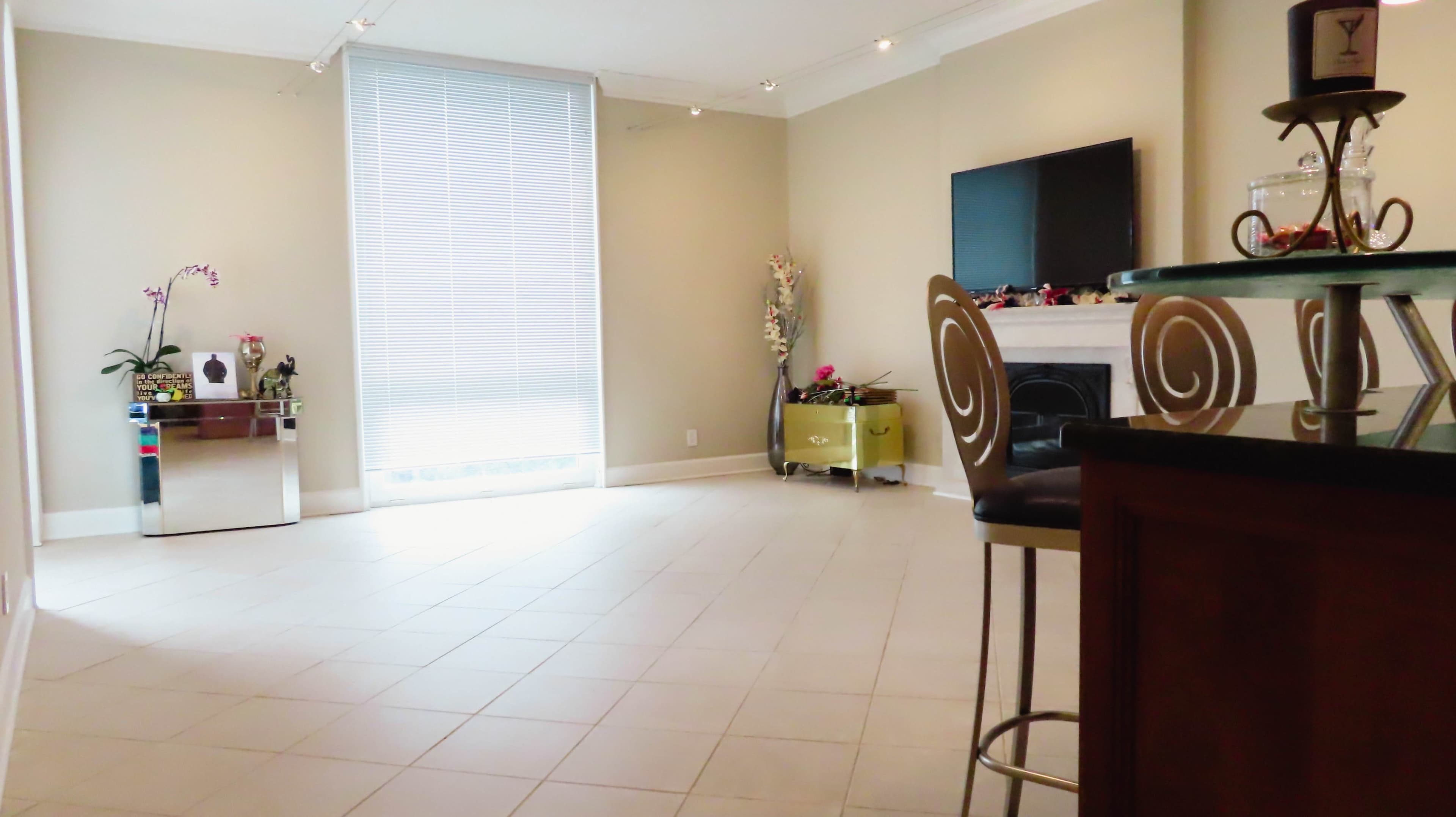 A modern living room features a flat-screen TV mounted on the wall, a decorative cabinet, and a small dining area with bar stools.