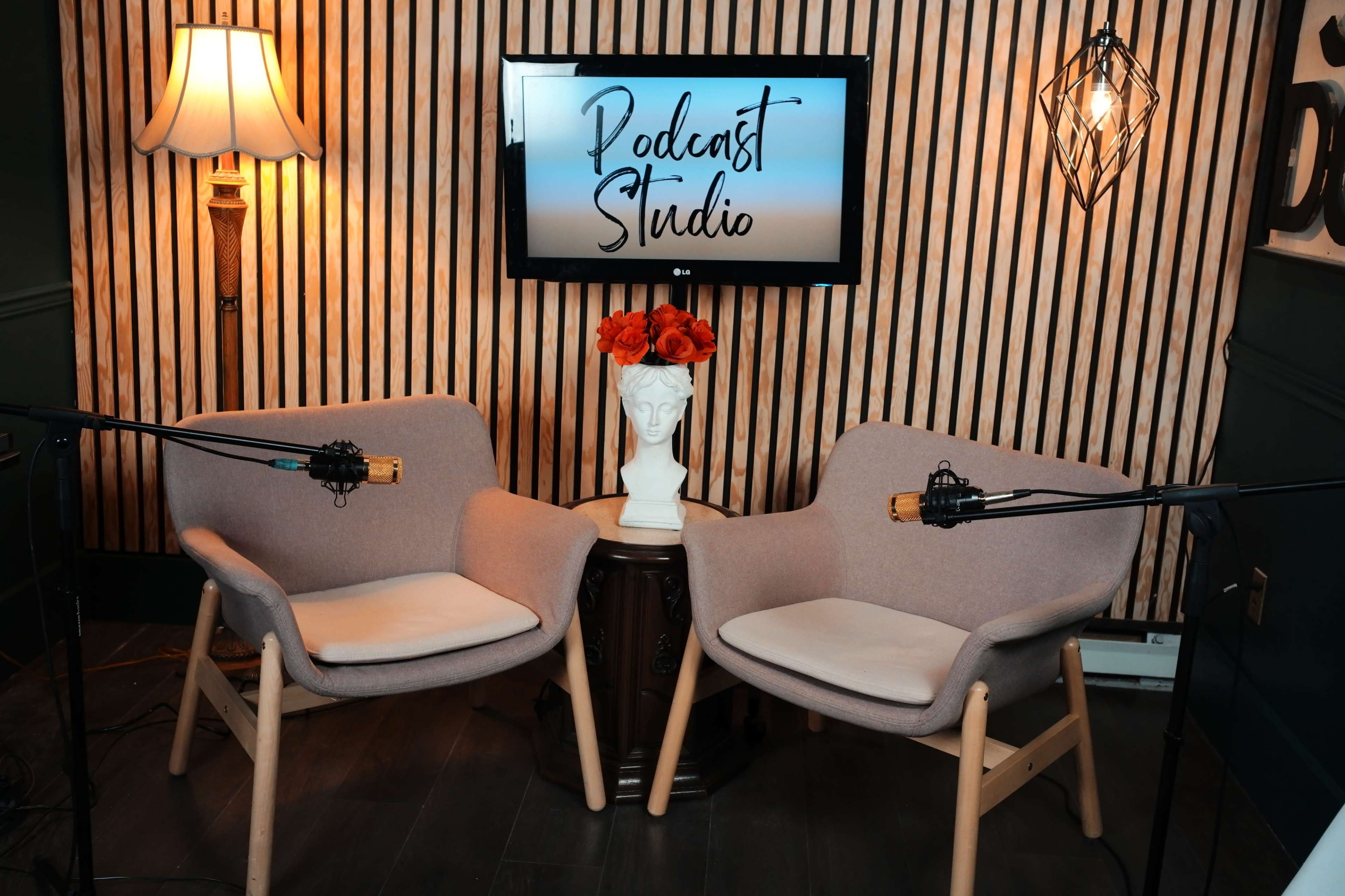 A podcast studio features two chairs, a small table with a sculpture, and microphones positioned in front of a screen displaying "Podcast Studio."