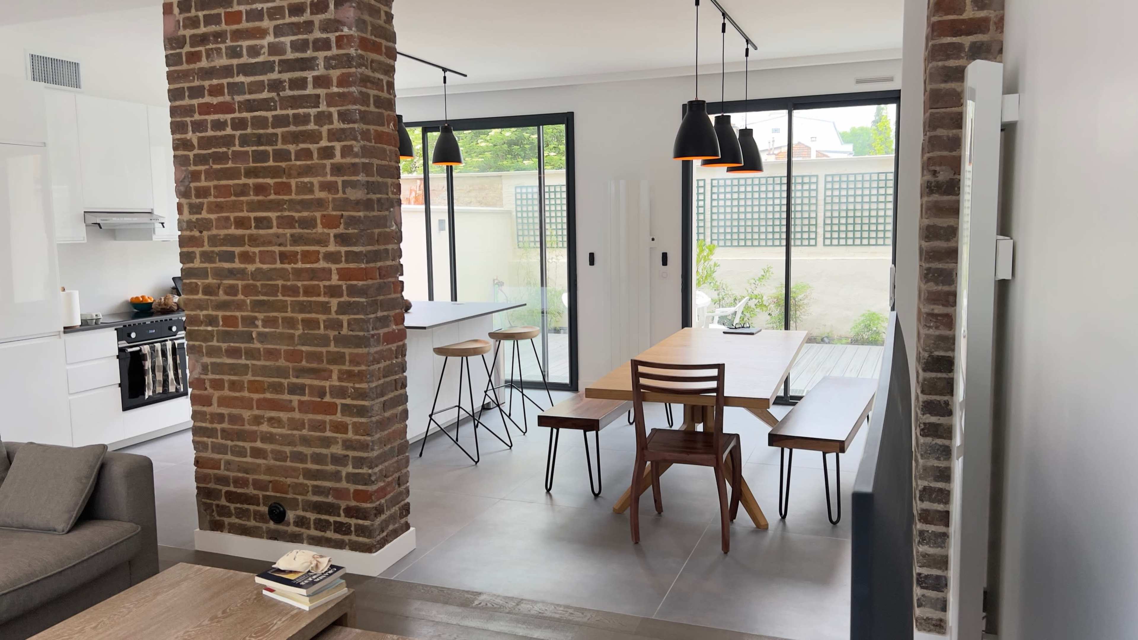 A modern open-plan living space with a brick column, a dining area with wooden furniture, and a kitchen visible in the background.