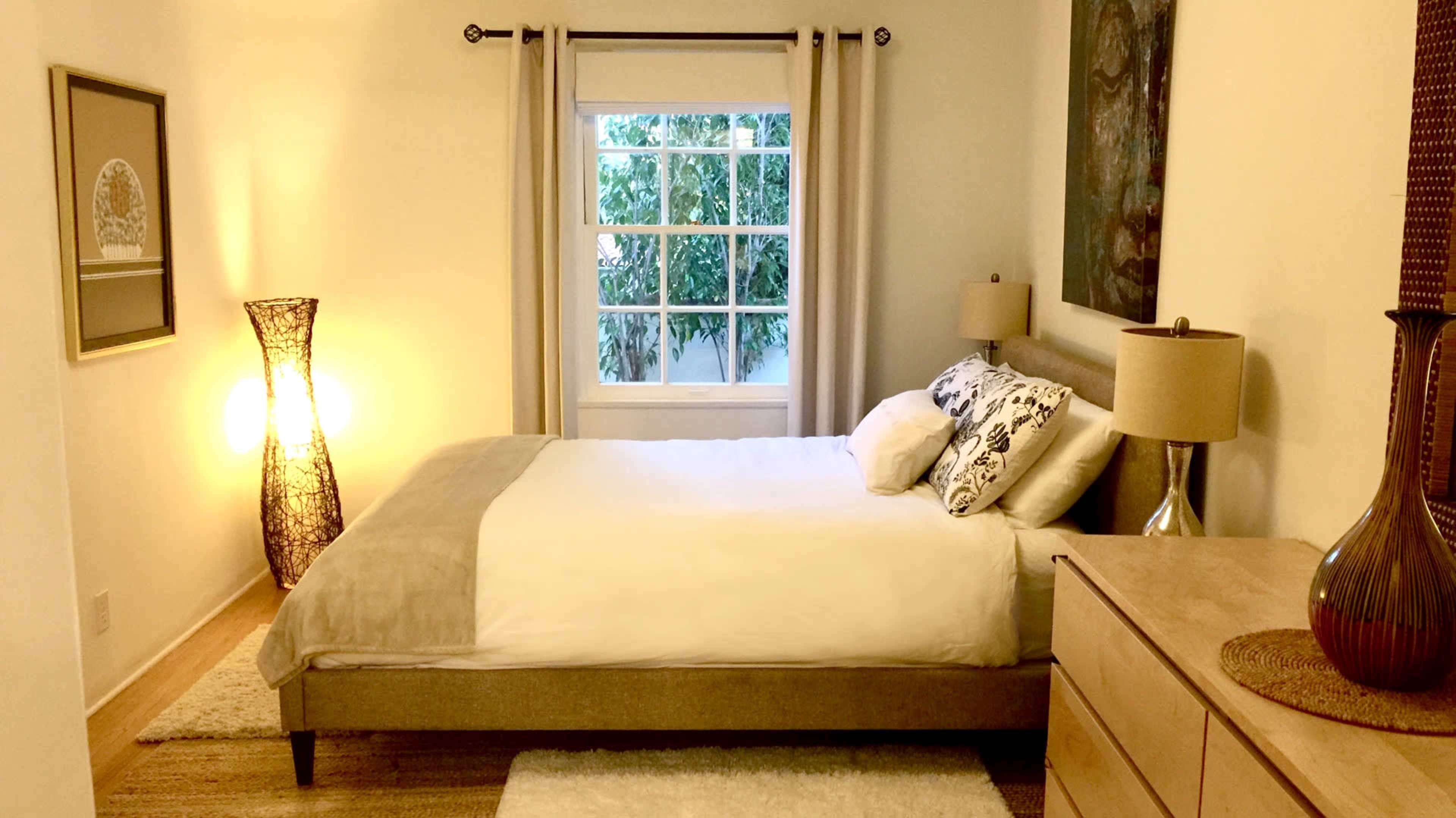 A well-lit bedroom features a bed with white bedding, a wooden dresser, and a large window with curtains overlooking greenery.