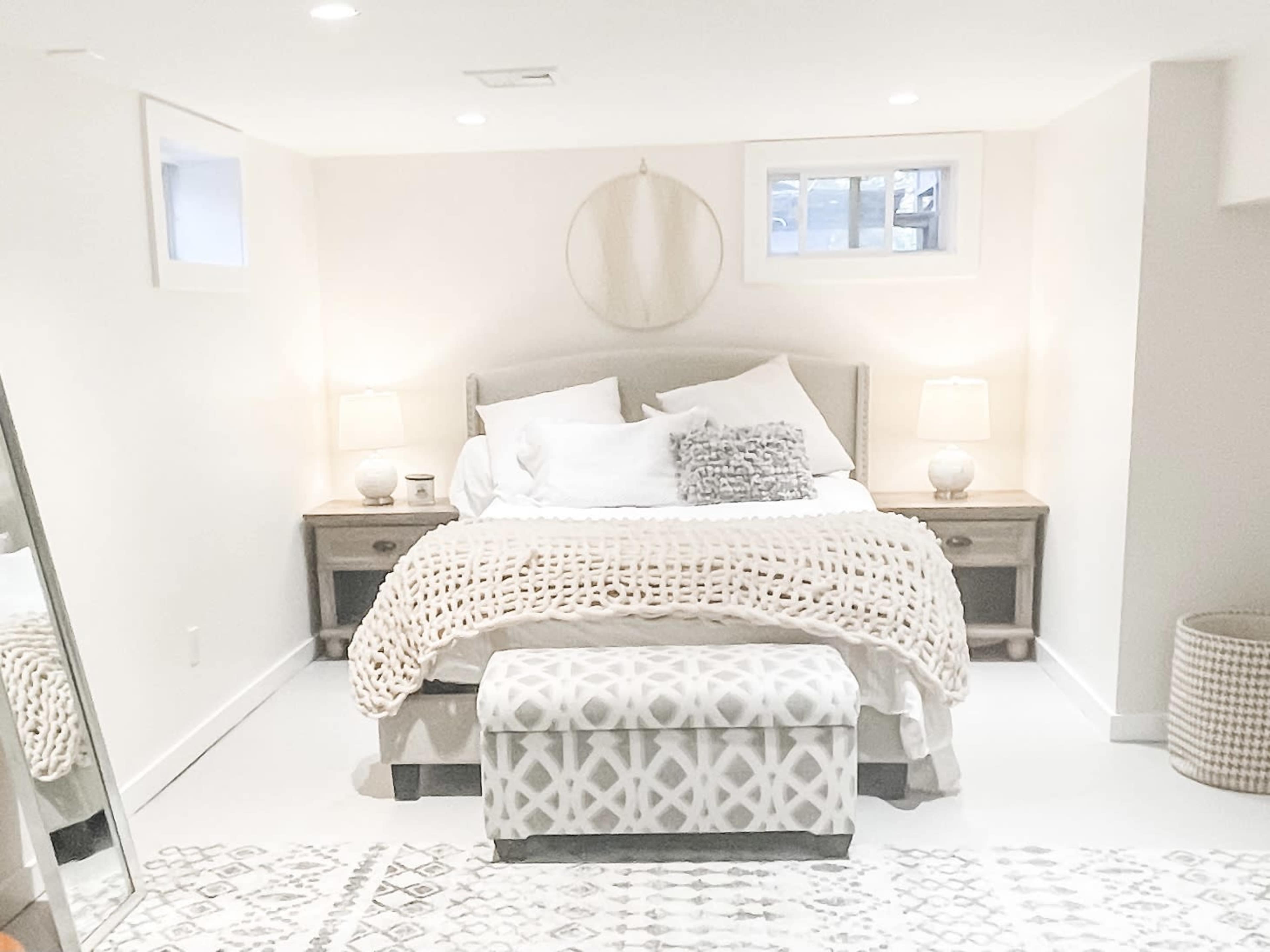 The image shows a softly lit bedroom featuring a bed with white linens, a patterned ottoman at the foot, and two bedside tables with lamps.
