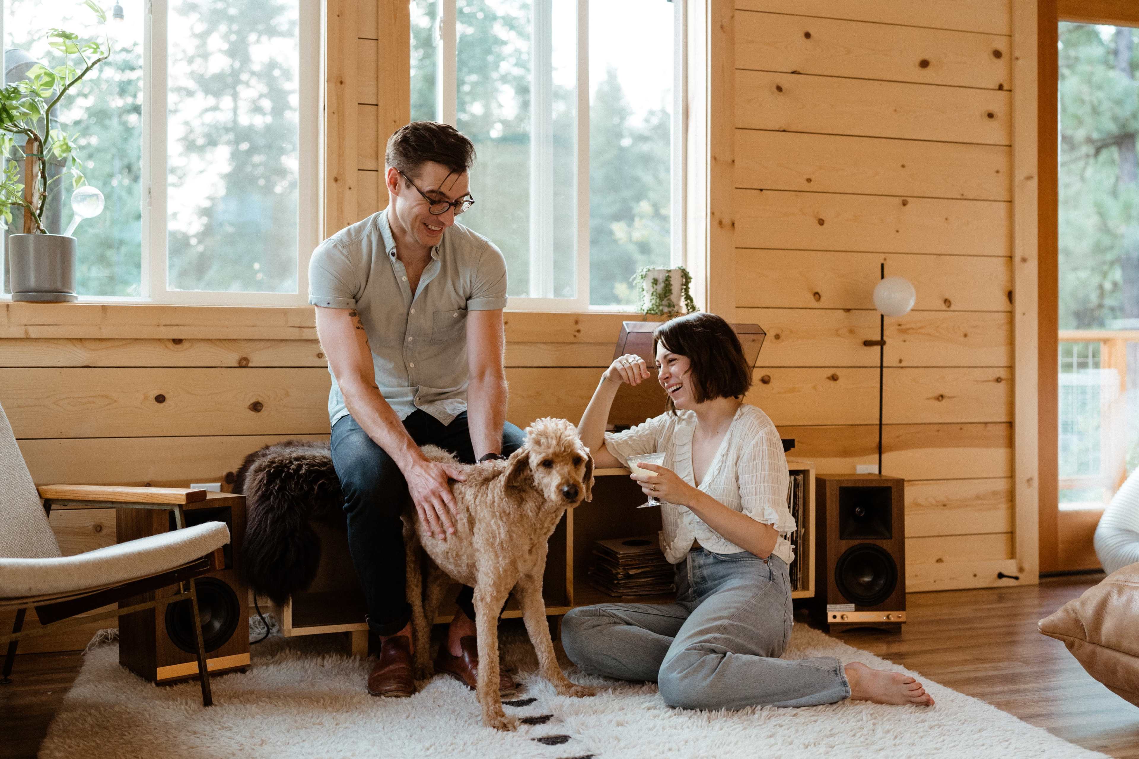 A man and a woman sit in a cozy, wooden room while interacting with a dog.