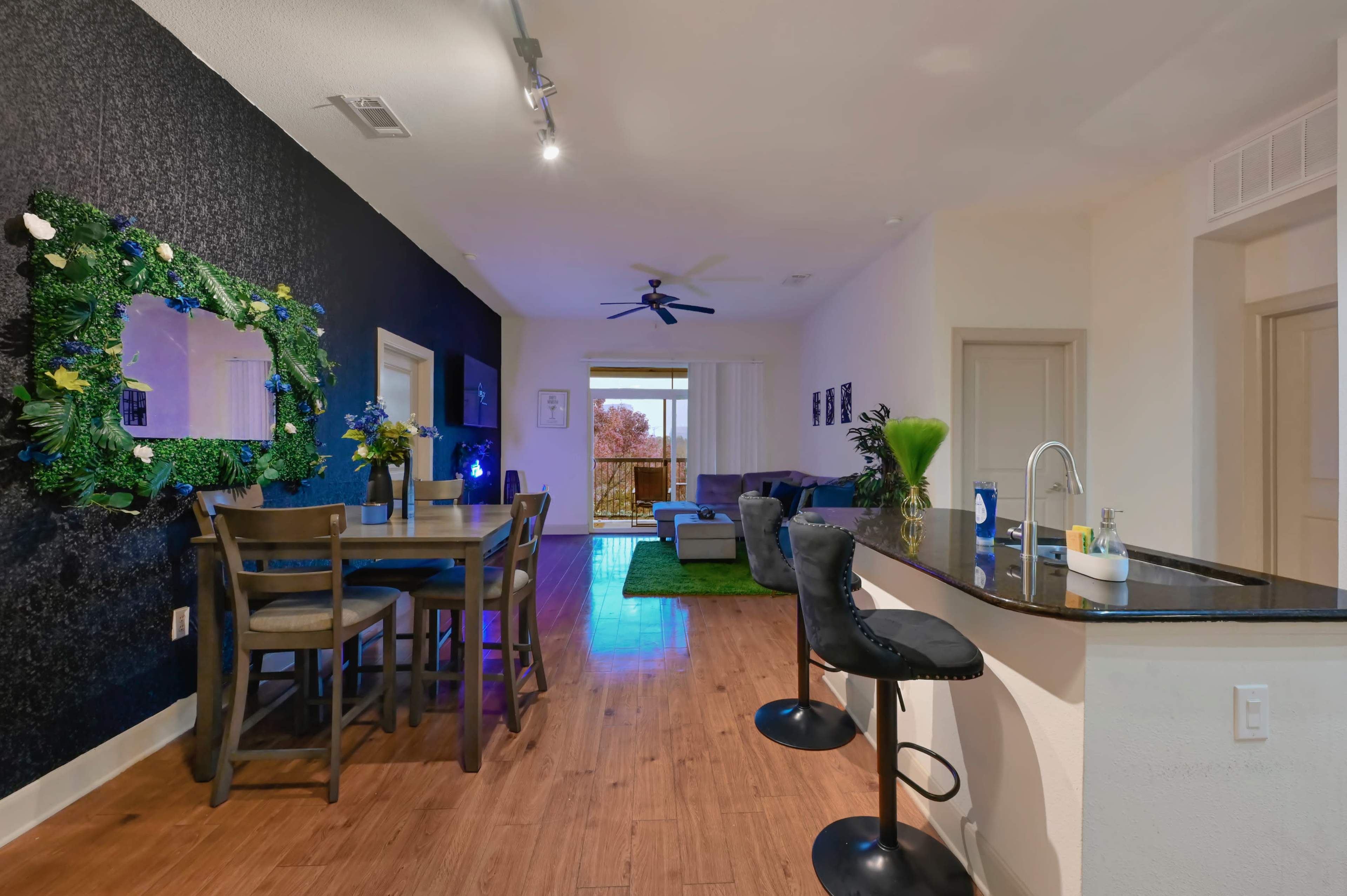 The image shows a modern living space featuring a dining area with a wooden table and chairs, a bar with stools, and a cozy lounge area with a couch and large window.