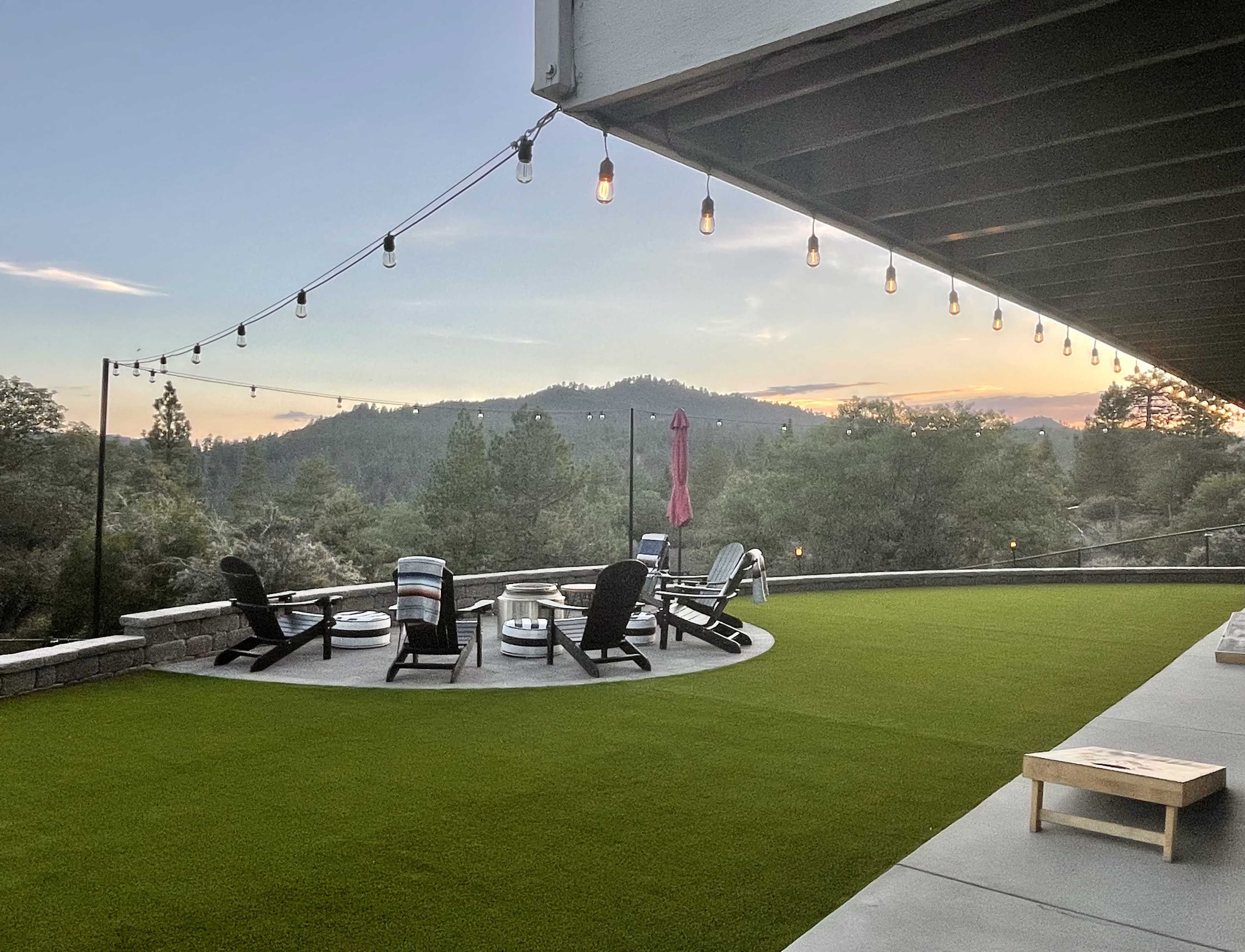 A spacious patio with circular seating, firepits, and string lights overlooks a mountainous landscape at sunset.