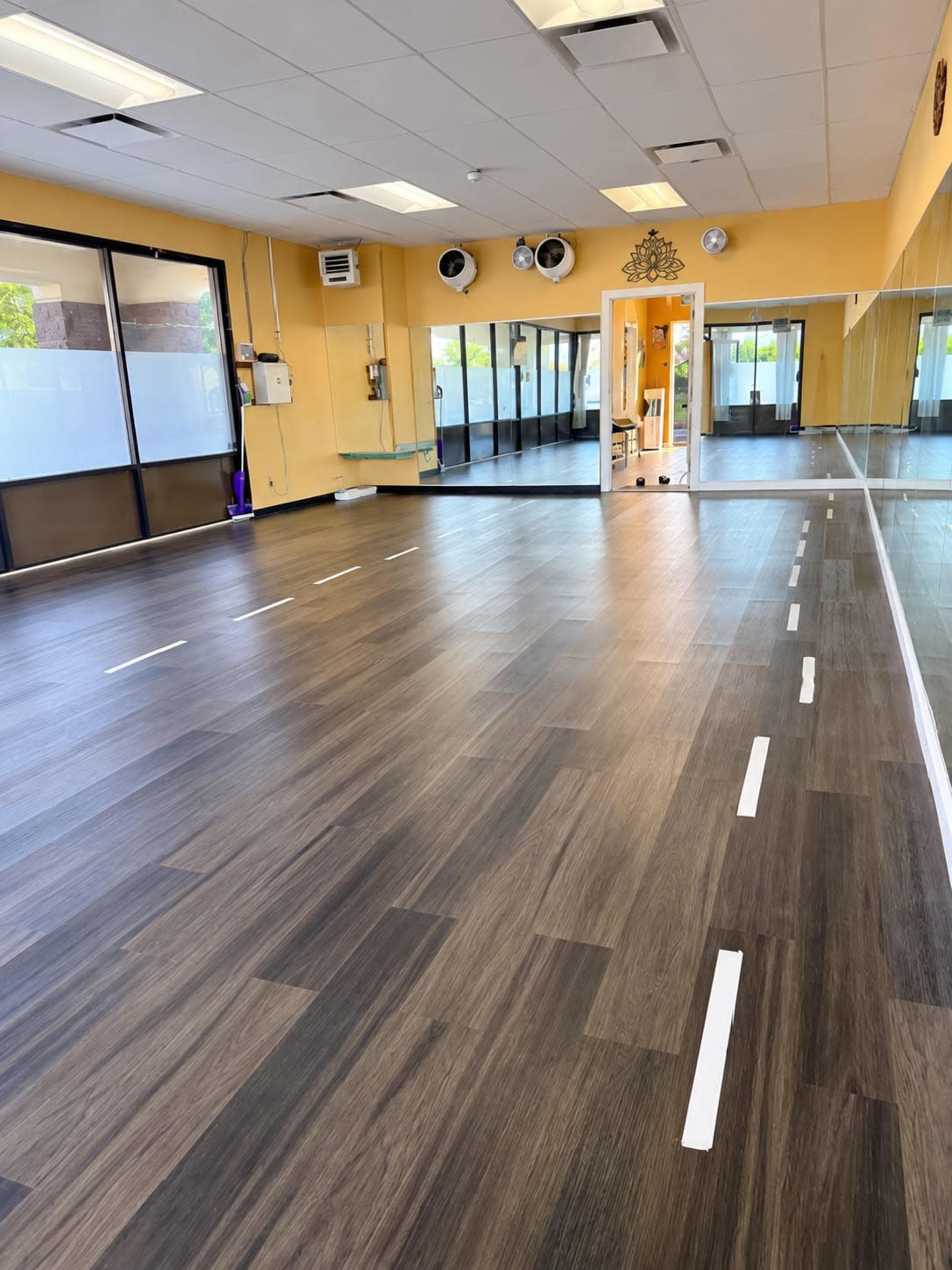 The image shows a spacious, well-lit room with a wooden floor, featuring white dashed lines on the floor and large mirrors along one wall.