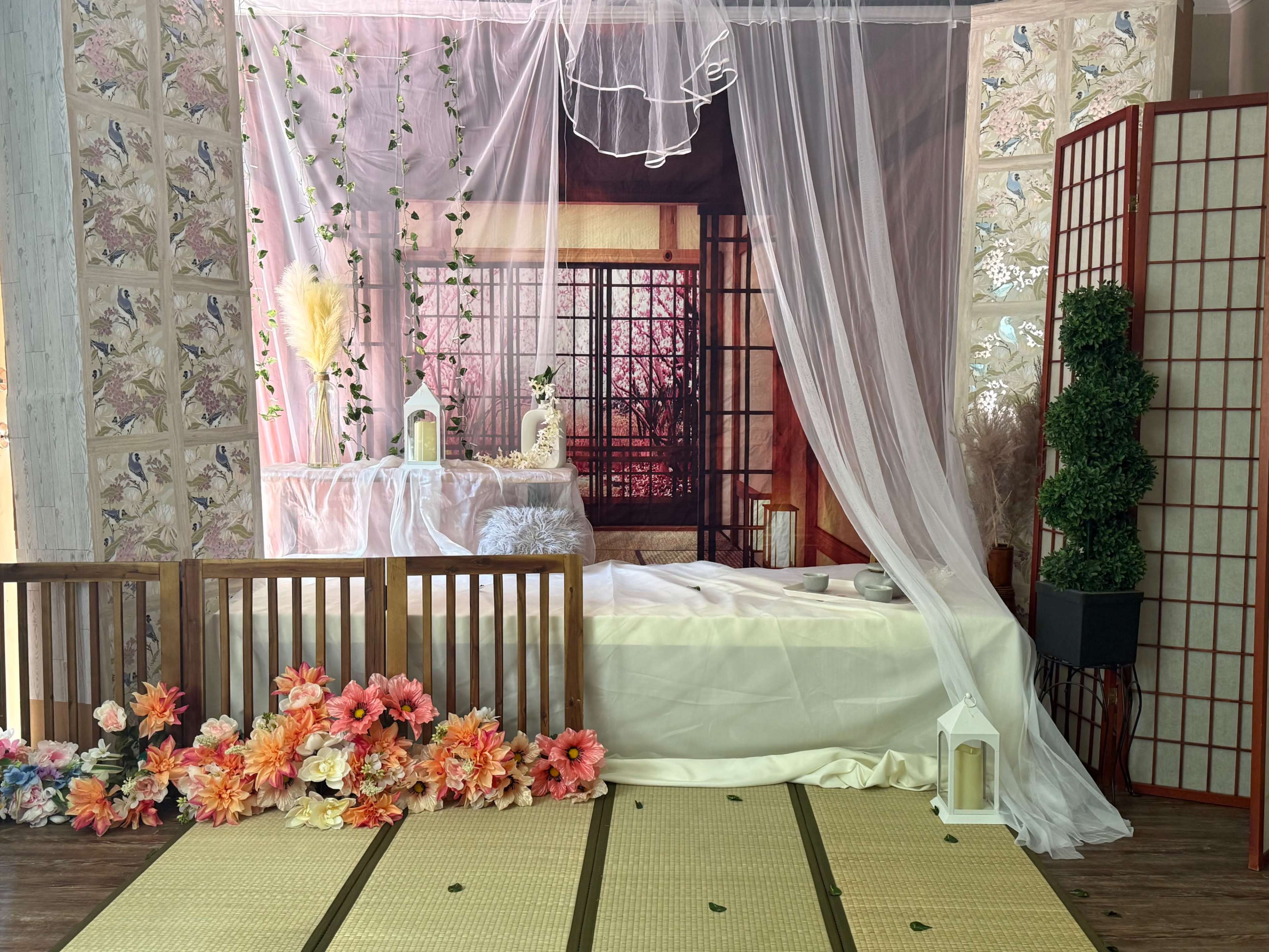 The scene features a decorated indoor space with a bed covered in white linens, framed by sheer drapes and surrounded by artificial flowers and decorative screens.