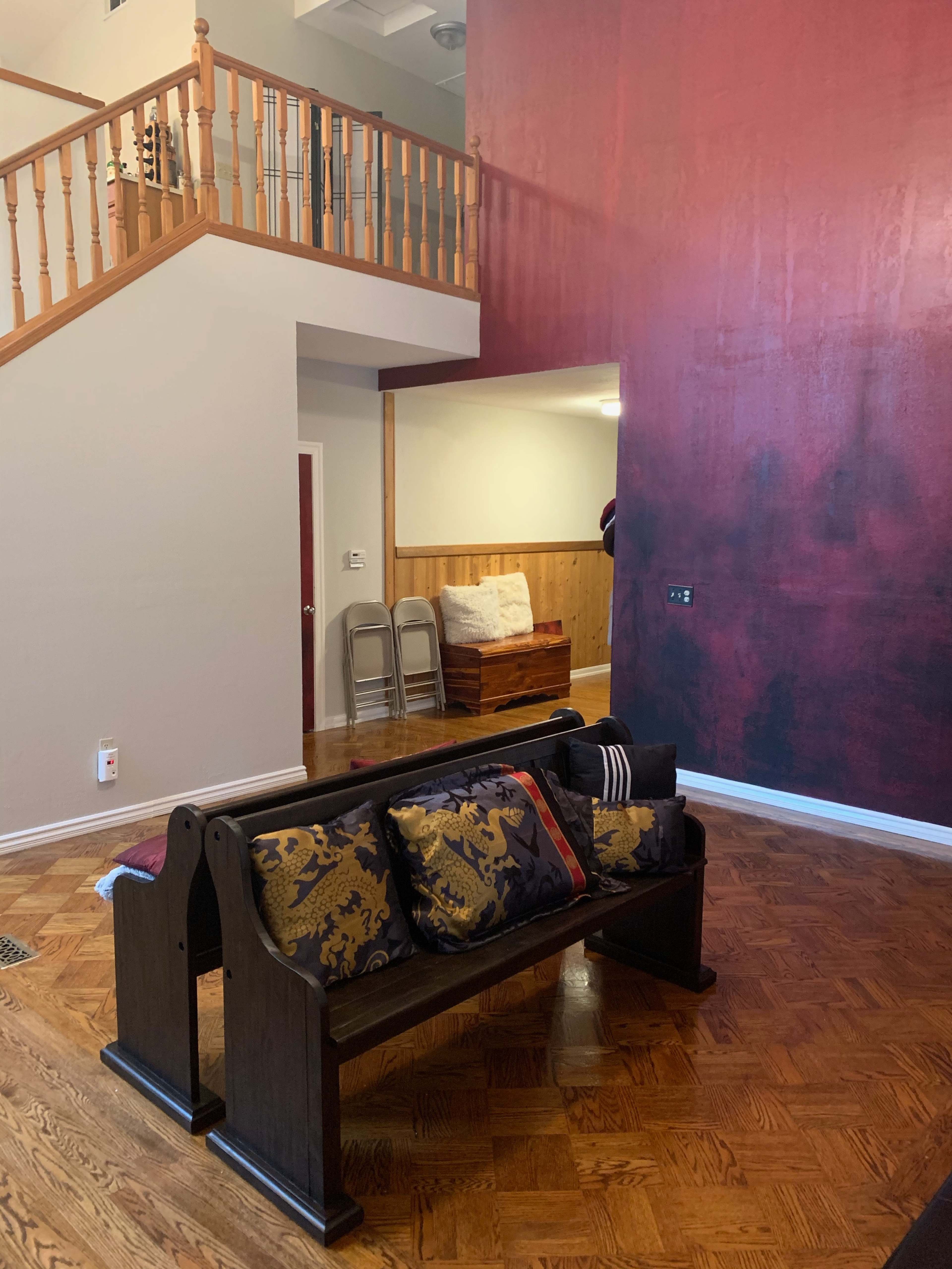 The image shows a living room with a dark red accent wall, a wooden bench adorned with patterned cushions, and a staircase in the background.