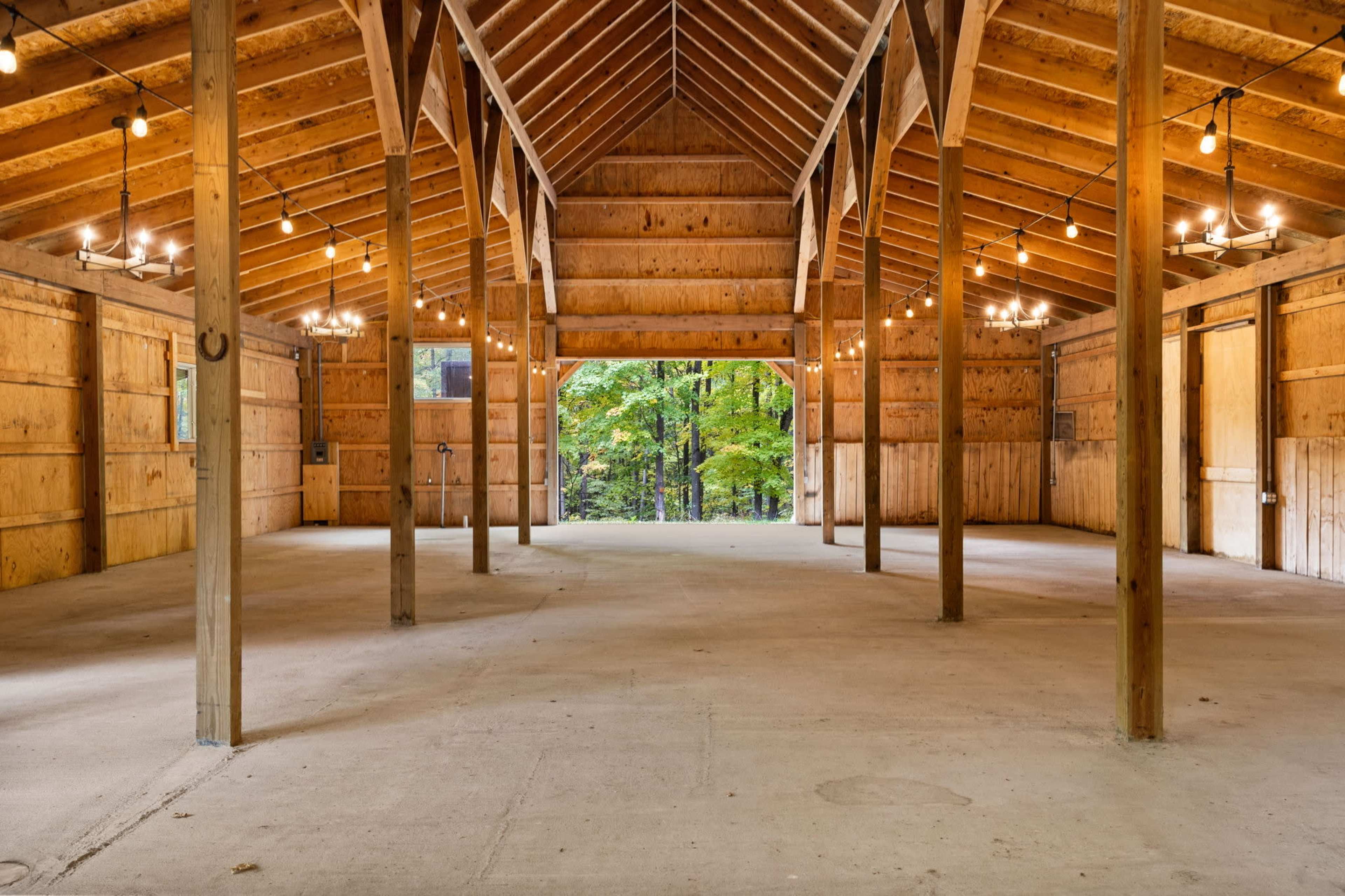 The image shows a large, empty wooden barn interior with exposed beams, chandeliers, and concrete flooring, surrounded by trees visible through the open doors.