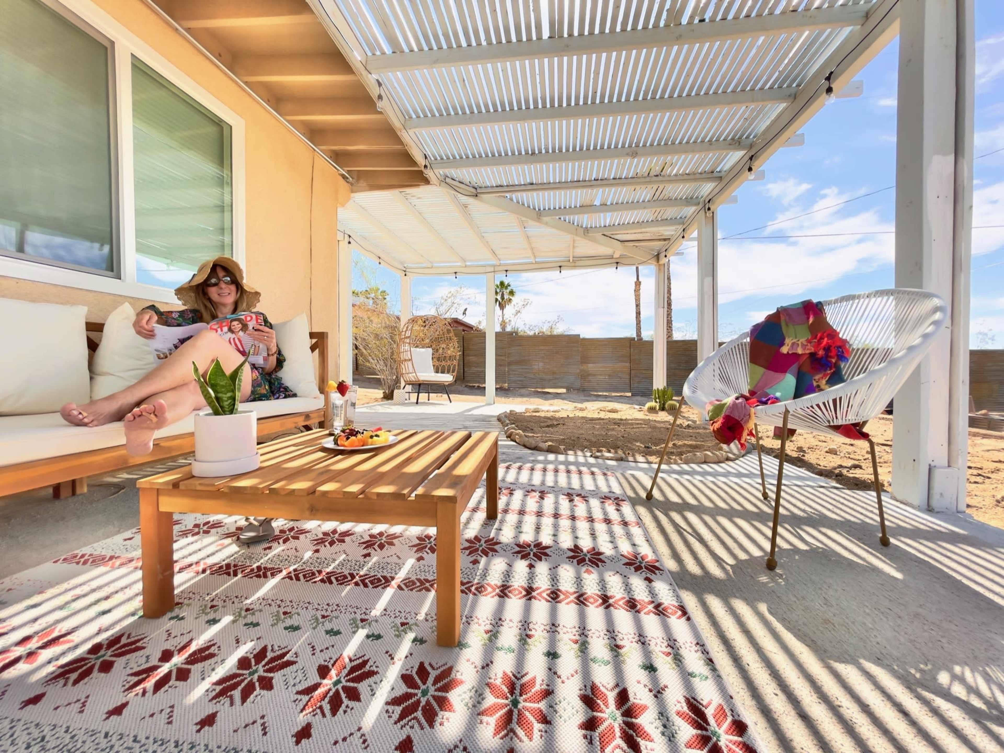 A woman relaxes on a couch under a shaded patio with a wooden table and decorative rug in a desert landscape.