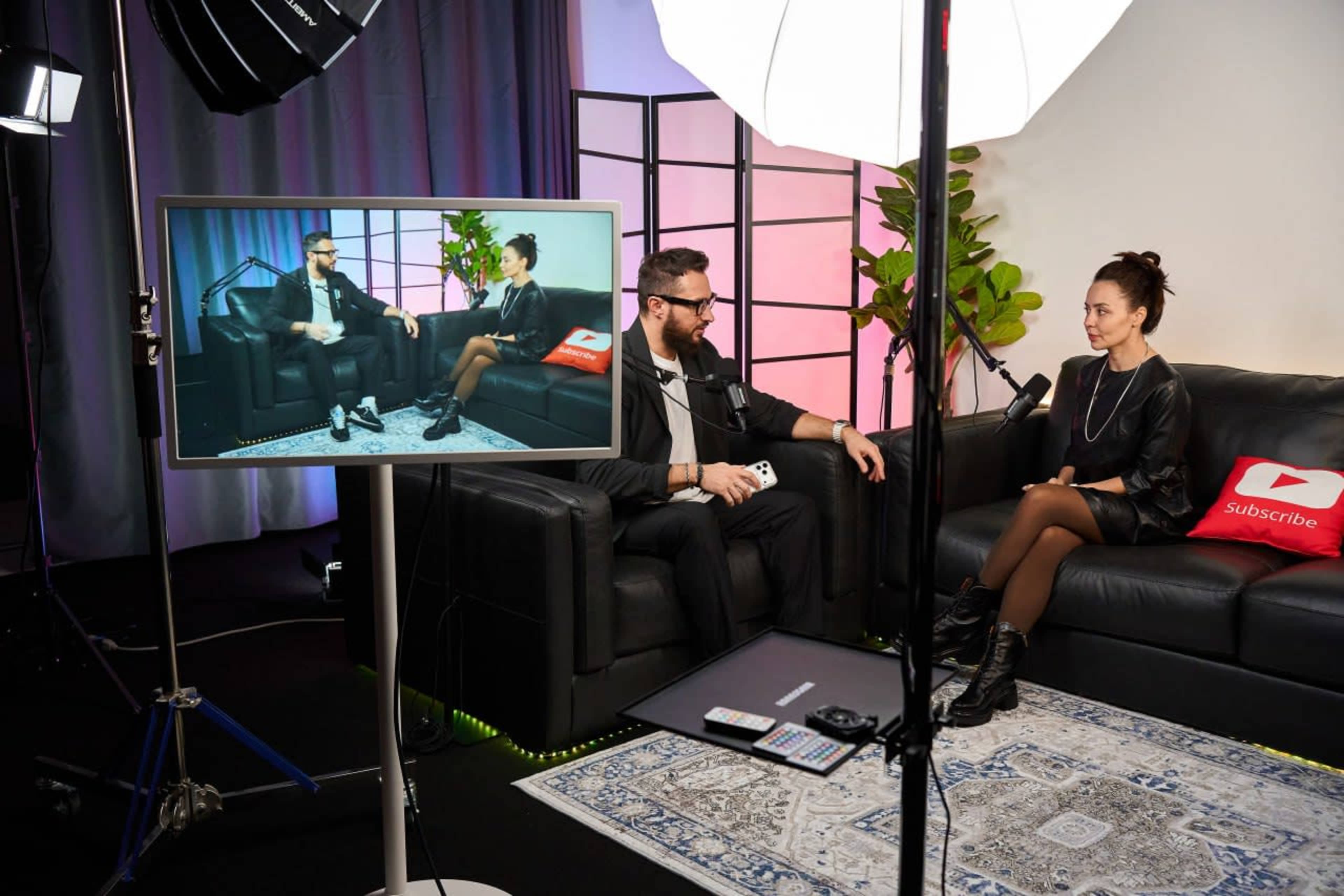 A man and a woman are seated on black sofas in a studio, with a camera capturing their conversation and a monitor displaying the scene.