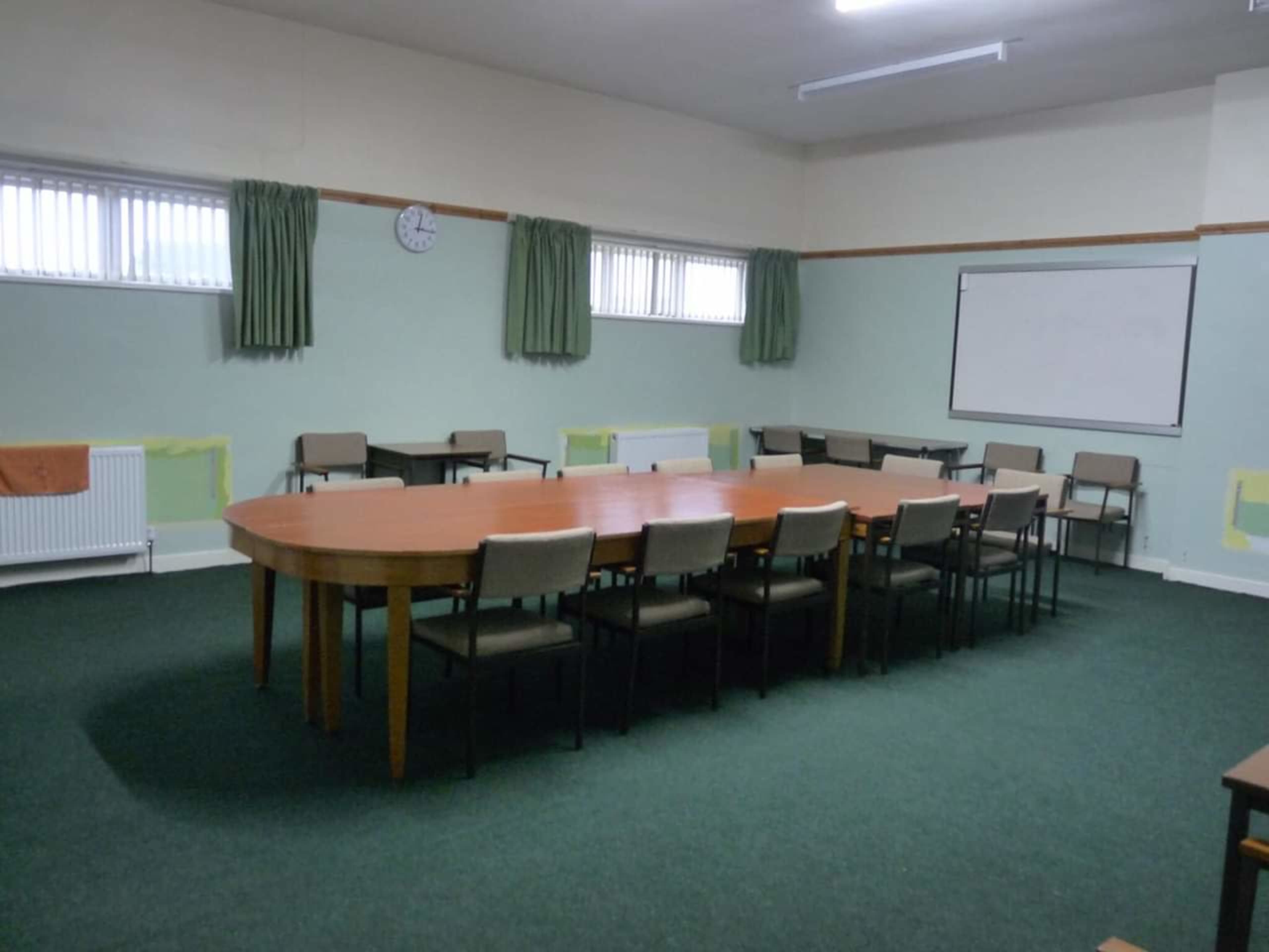 A large, oval table surrounded by chairs is set in an empty meeting room with green carpeting and small windows.