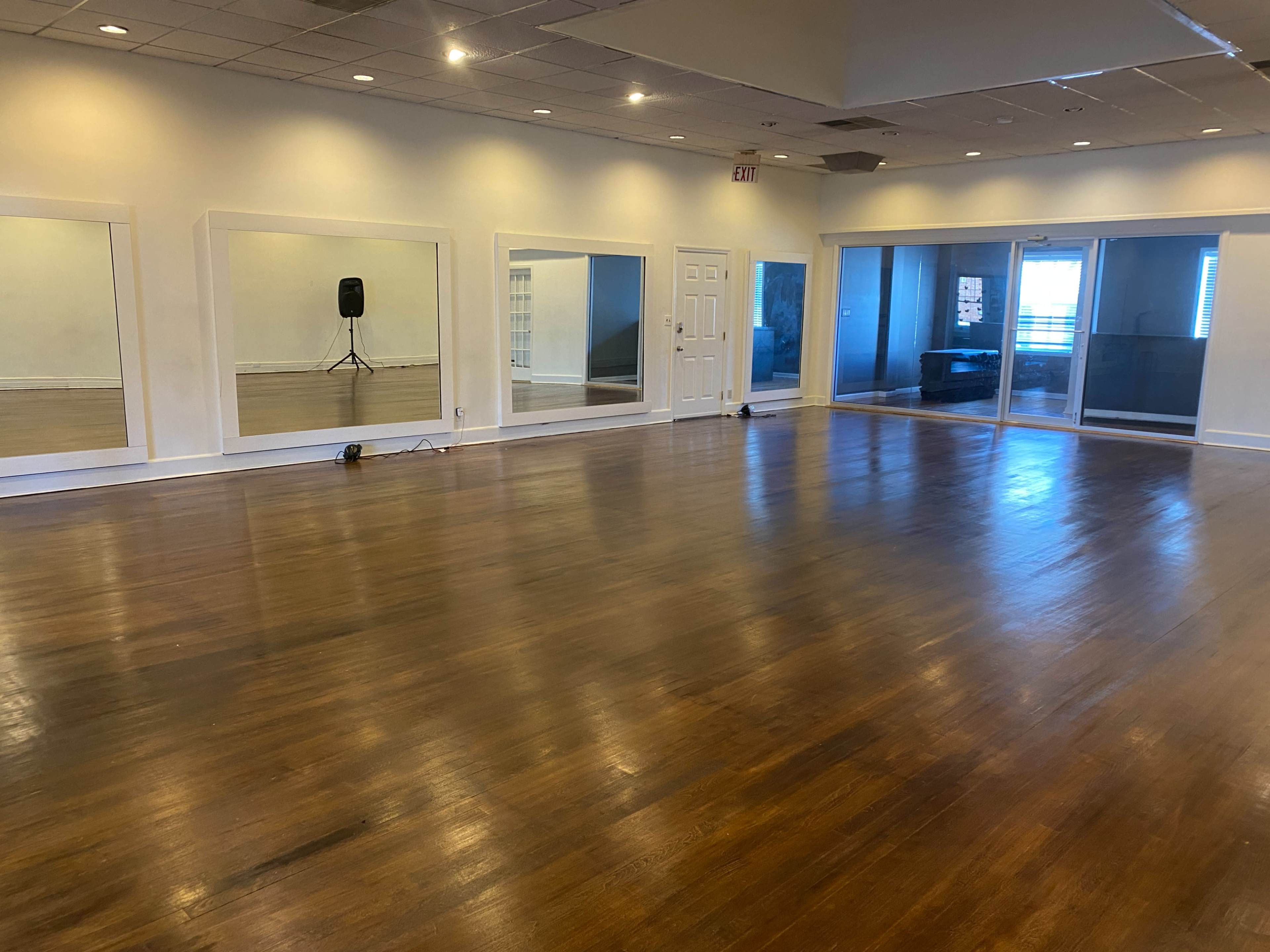 The image shows a spacious, empty room with polished wooden floors, large mirrors on one wall, and glass doors leading to an outdoor area.