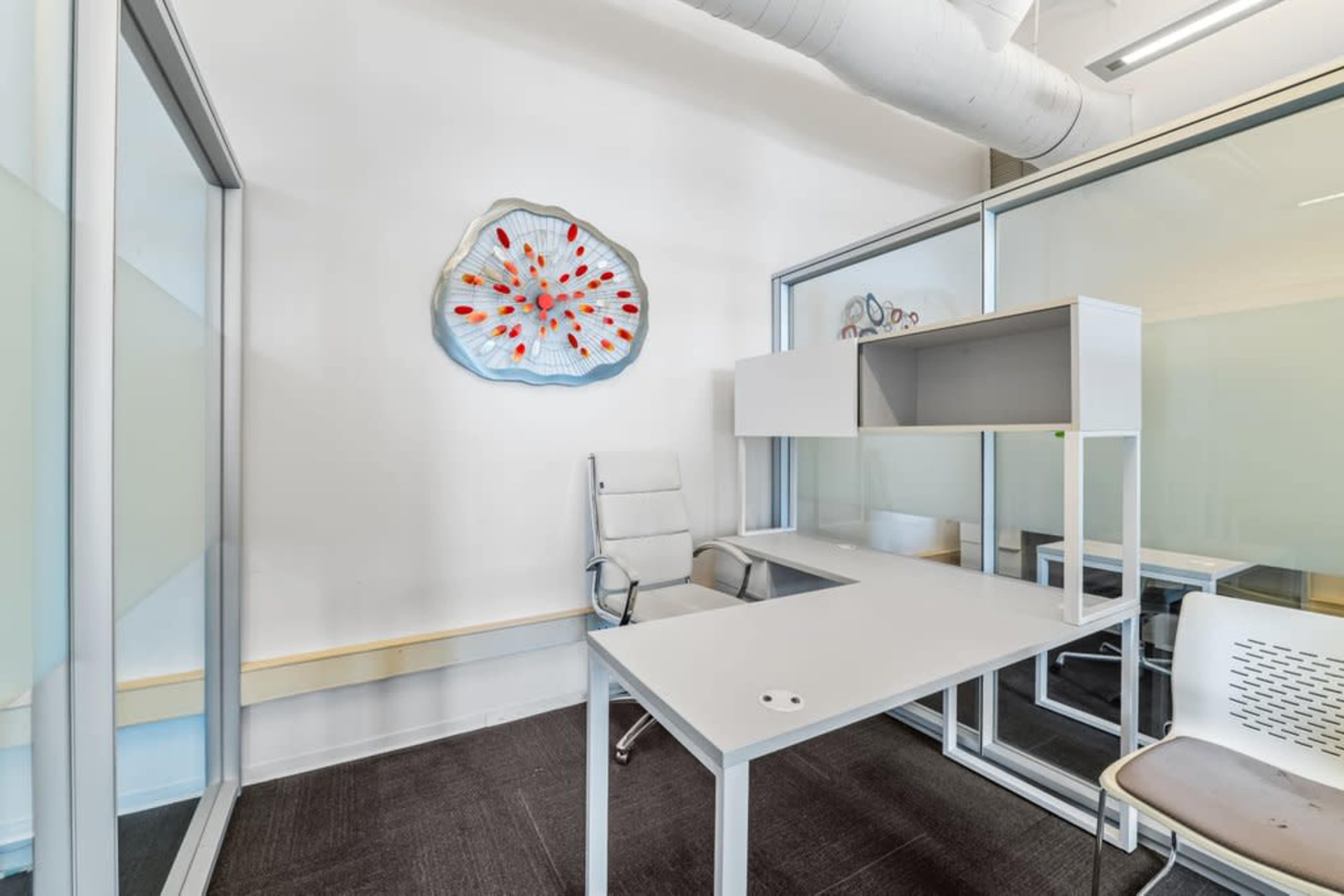 The image shows a minimalist office space with a white desk, a clear chair, and an abstract wall art piece.