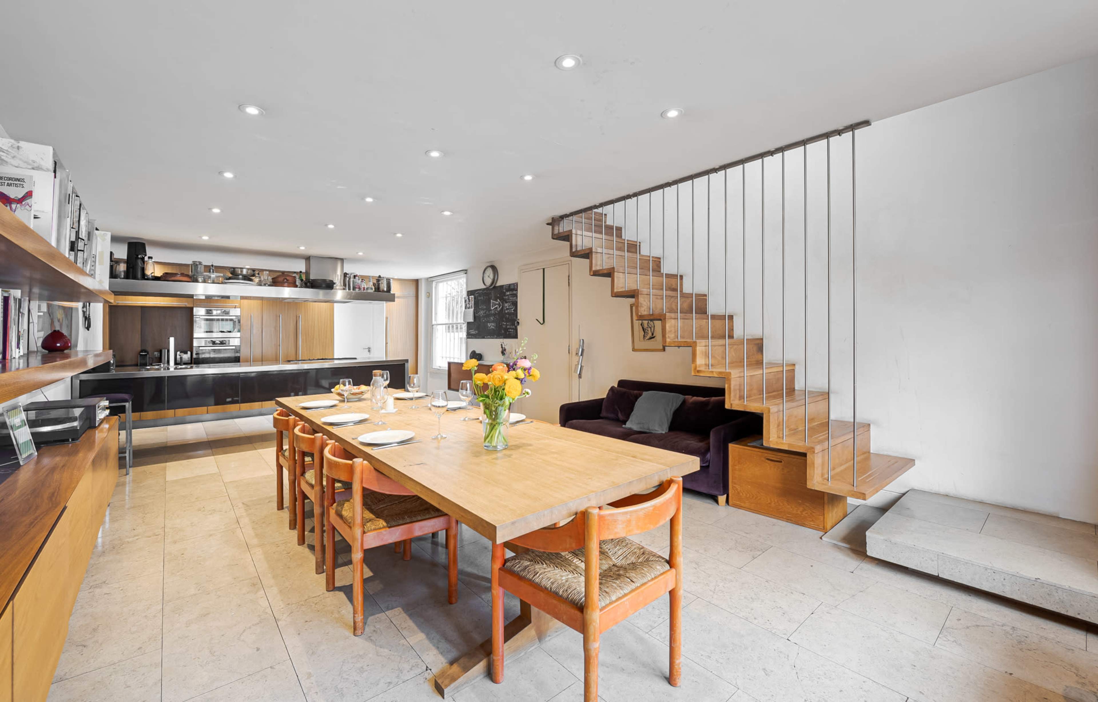 A modern kitchen and dining area features a wooden table with seating, a sleek kitchen counter, and a staircase leading to an upper level.