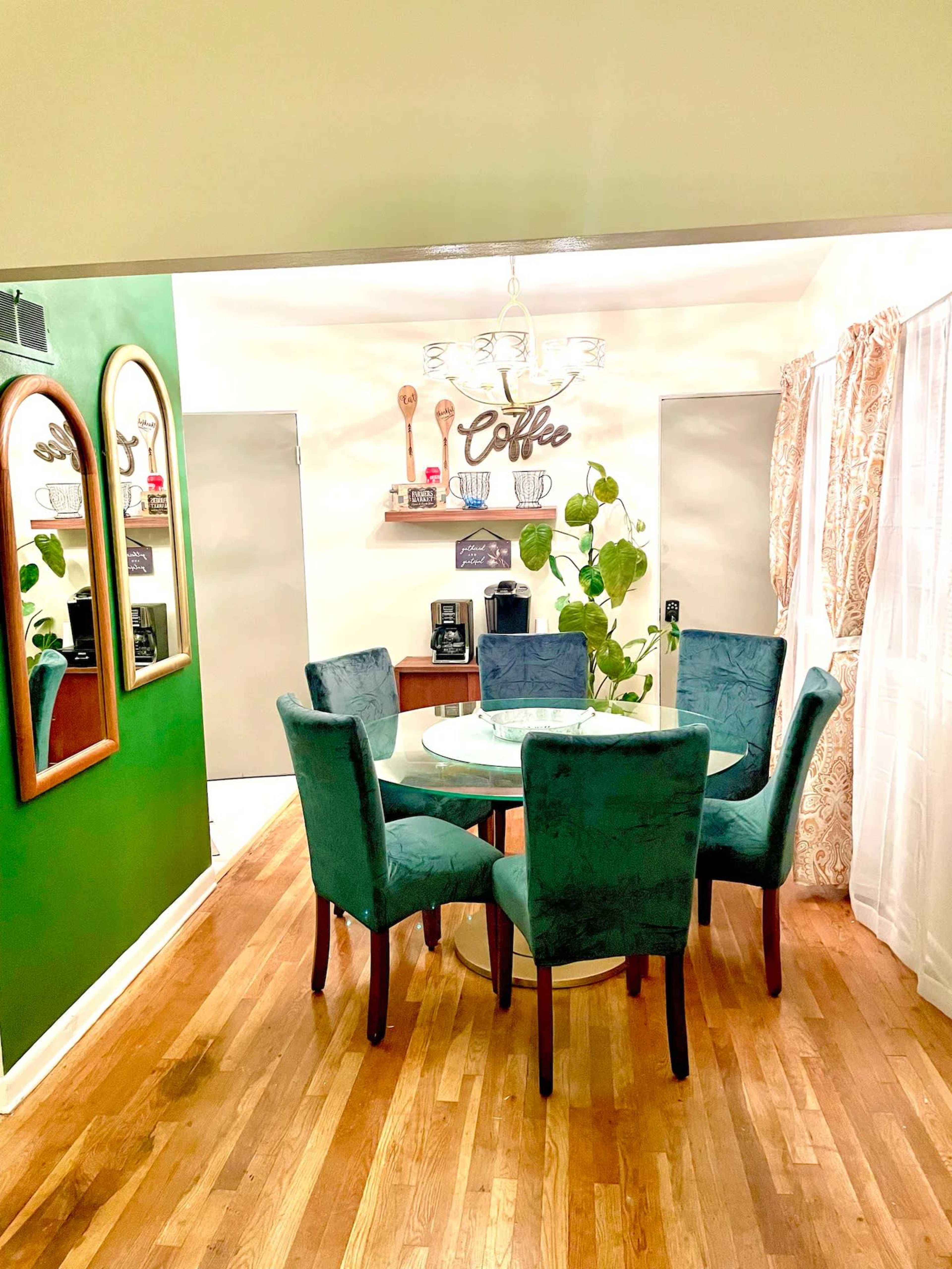A dining area with a round glass table surrounded by six green chairs, a coffee station, and decorative wall shelves in a room with green and white walls.