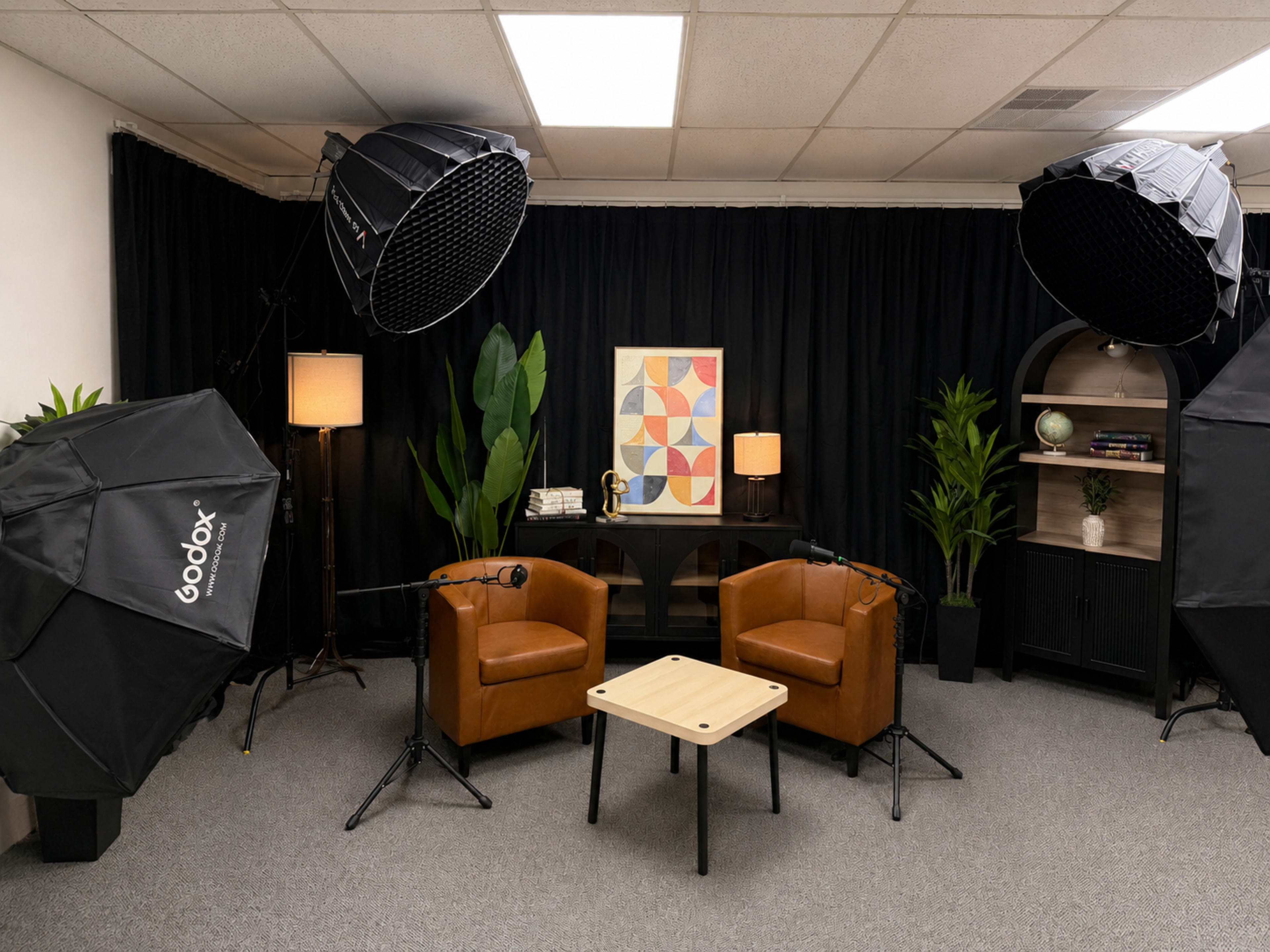 The image shows a well-lit interview setup with two brown chairs, a small table, and studio lighting equipment in a room with a black curtain backdrop.