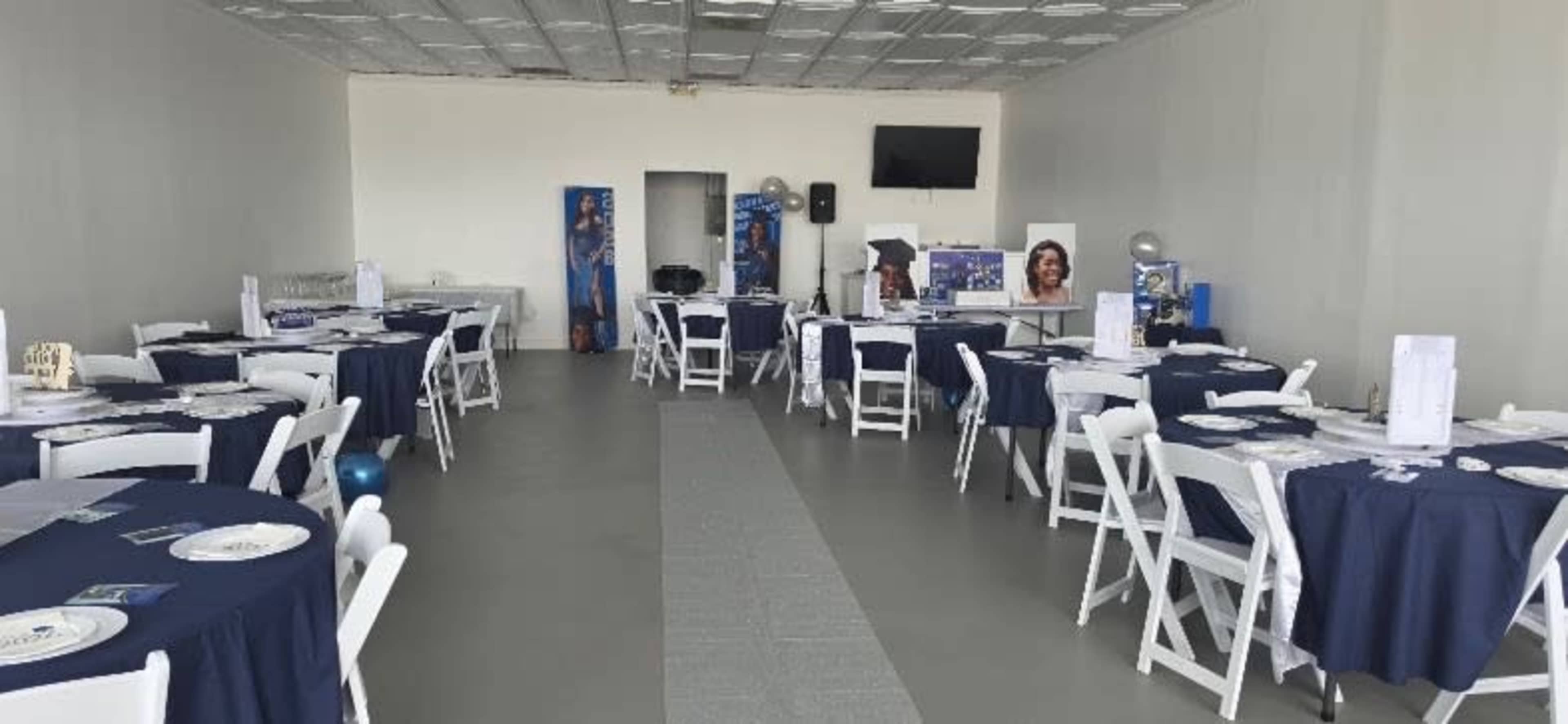 The interior of a large room is set up for an event, featuring multiple tables covered with dark blue tablecloths and white chairs arranged neatly, along with a presentation area in the background.