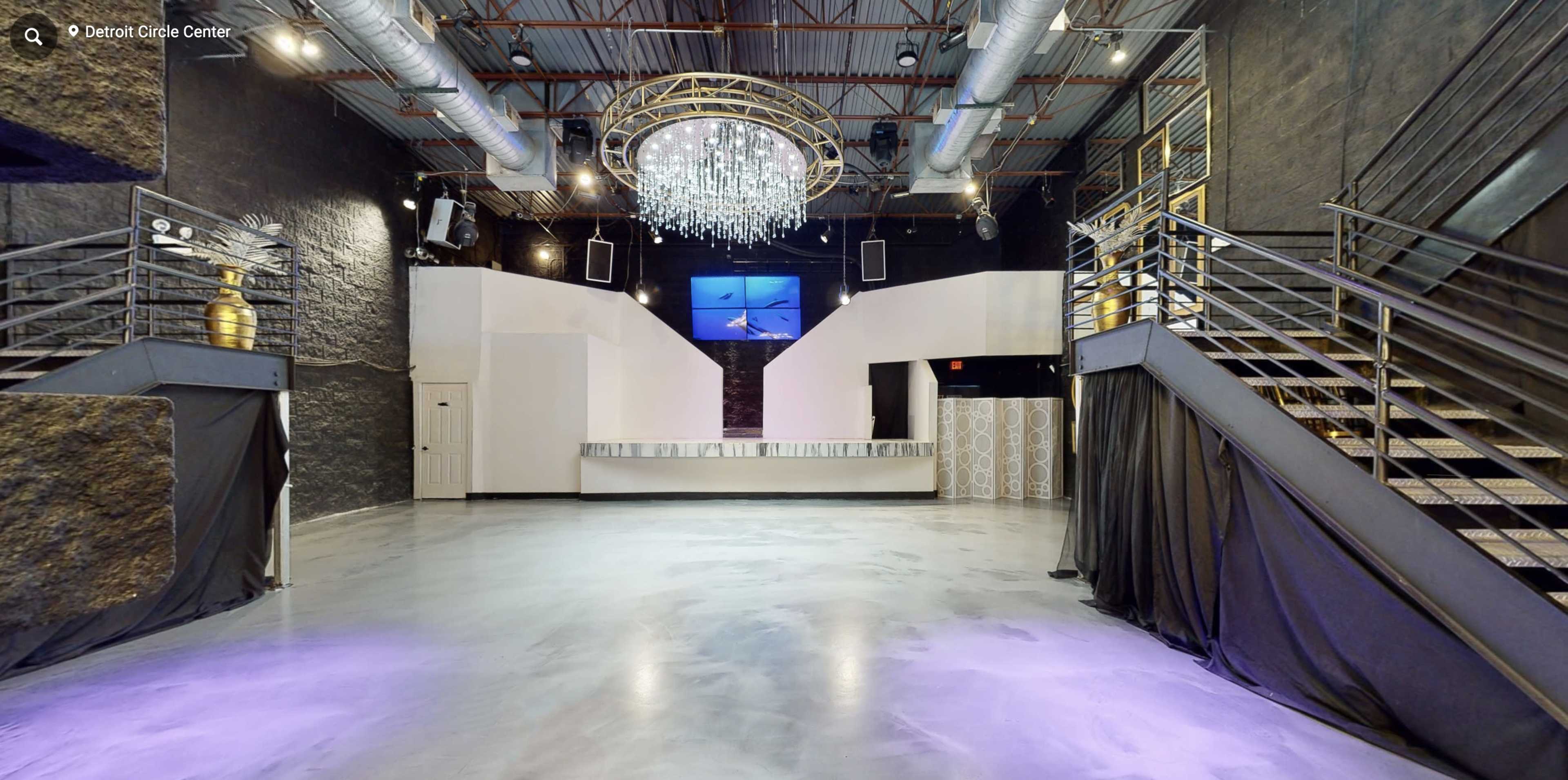 A spacious event venue with a central chandelier, a bar area at the back, and two staircases leading to an upper level.