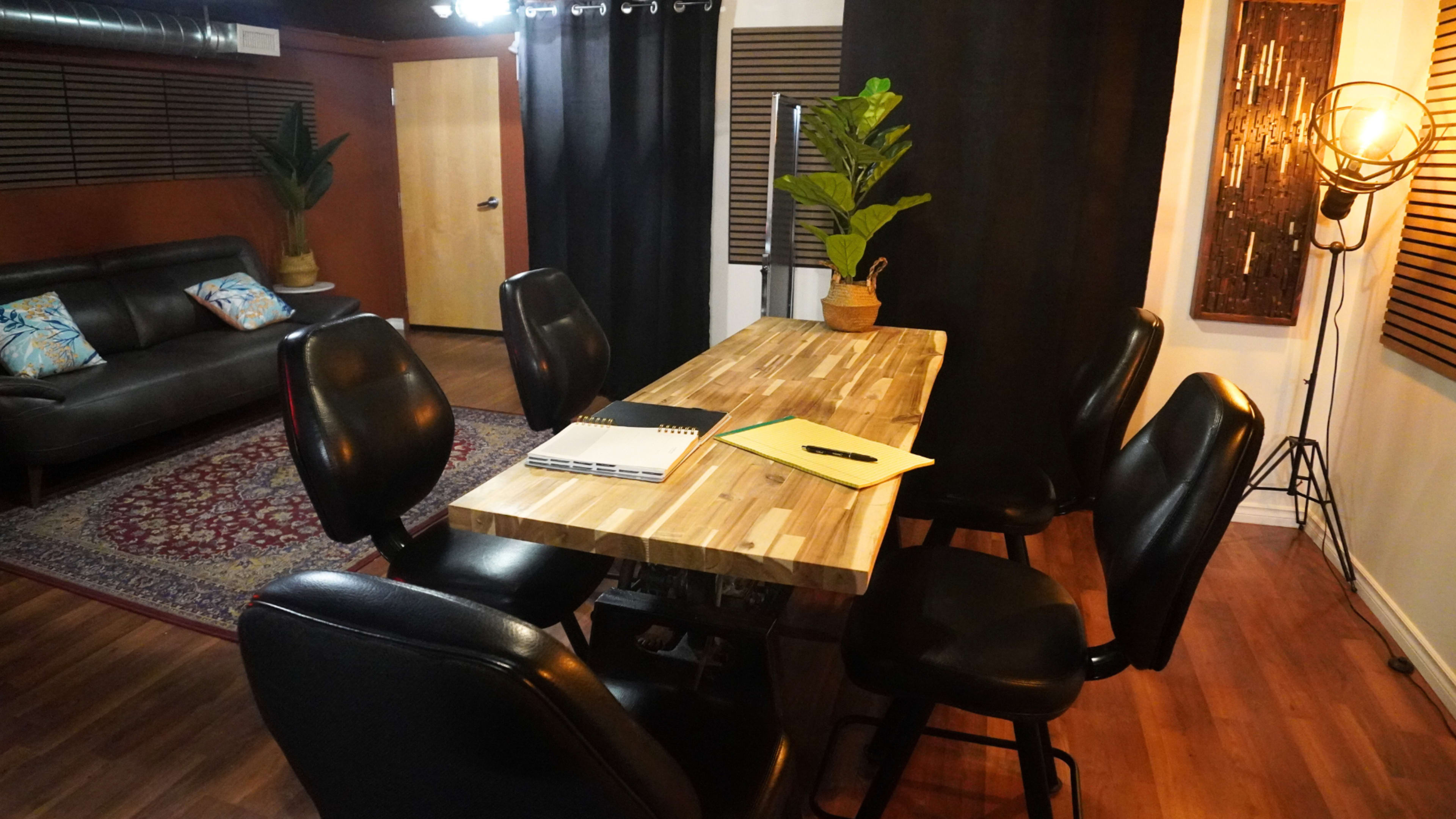 The image shows a modern meeting room featuring a wooden table surrounded by six black chairs, with a plant in a pot and a couch in the background.