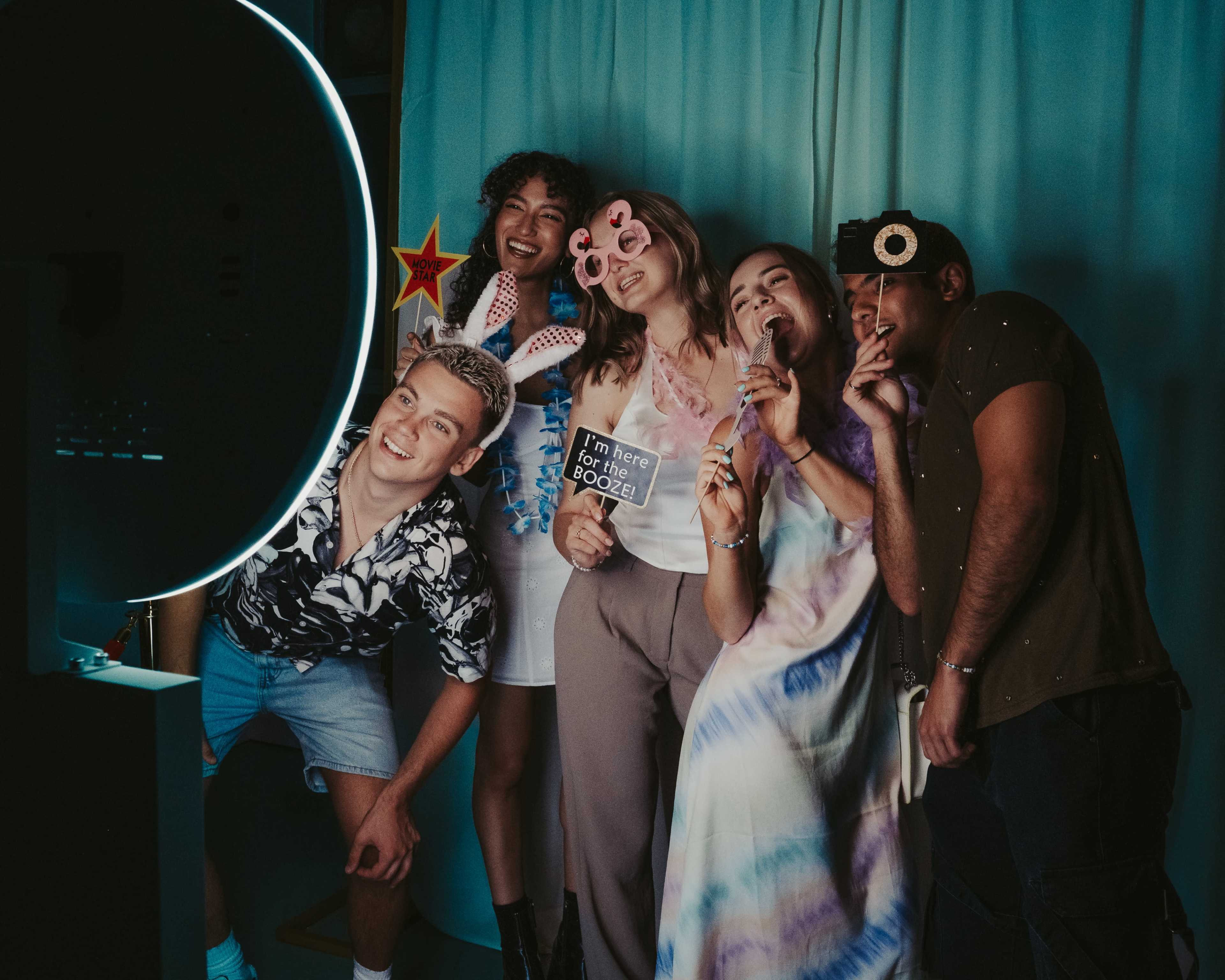 A group of five friends poses playfully in front of a circular light, holding props and making fun gestures against a backdrop of blue fabric.