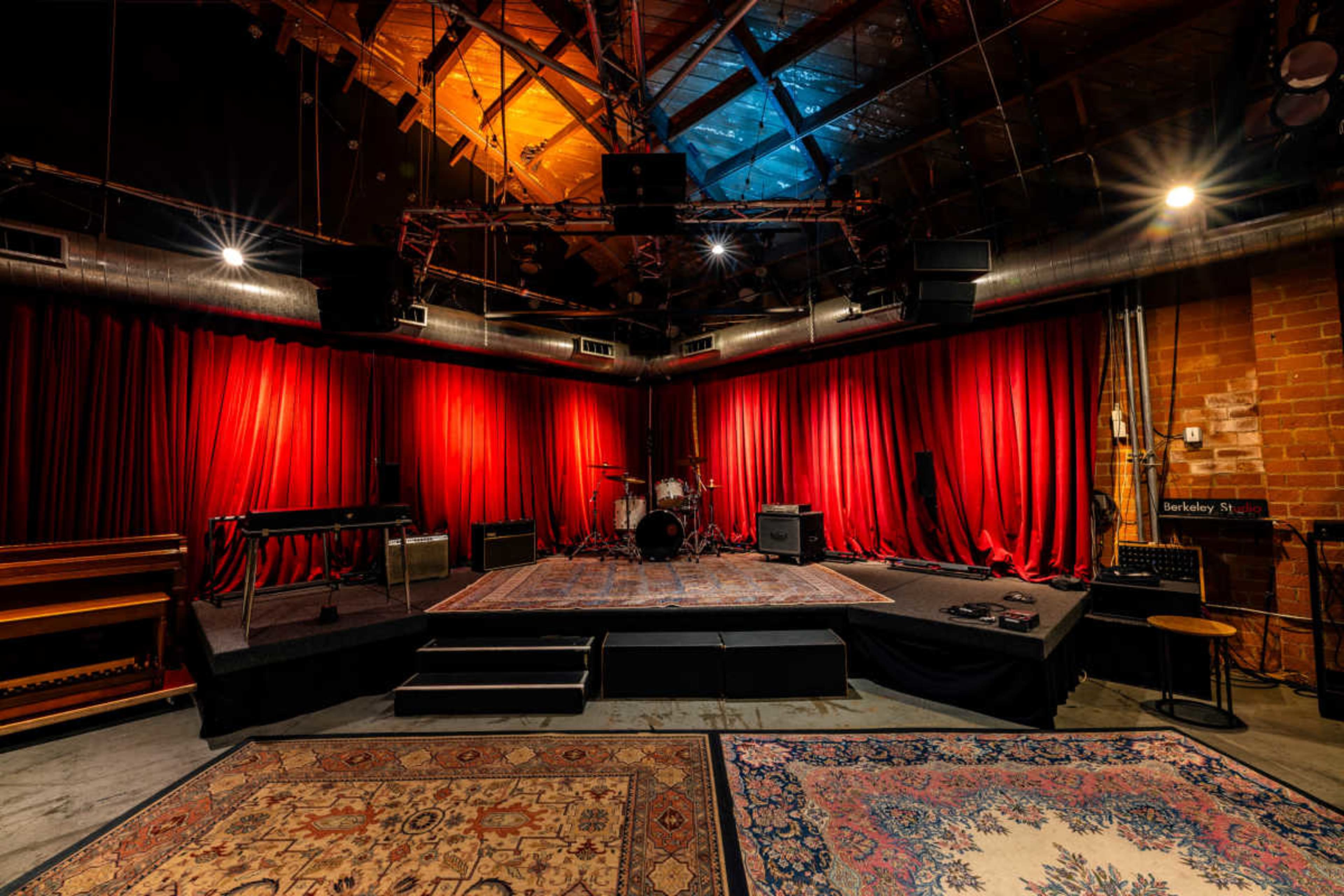 The image shows a music performance stage with red curtains, a drum set, amplifiers, and rugs on the floor, all within a dimly lit room.