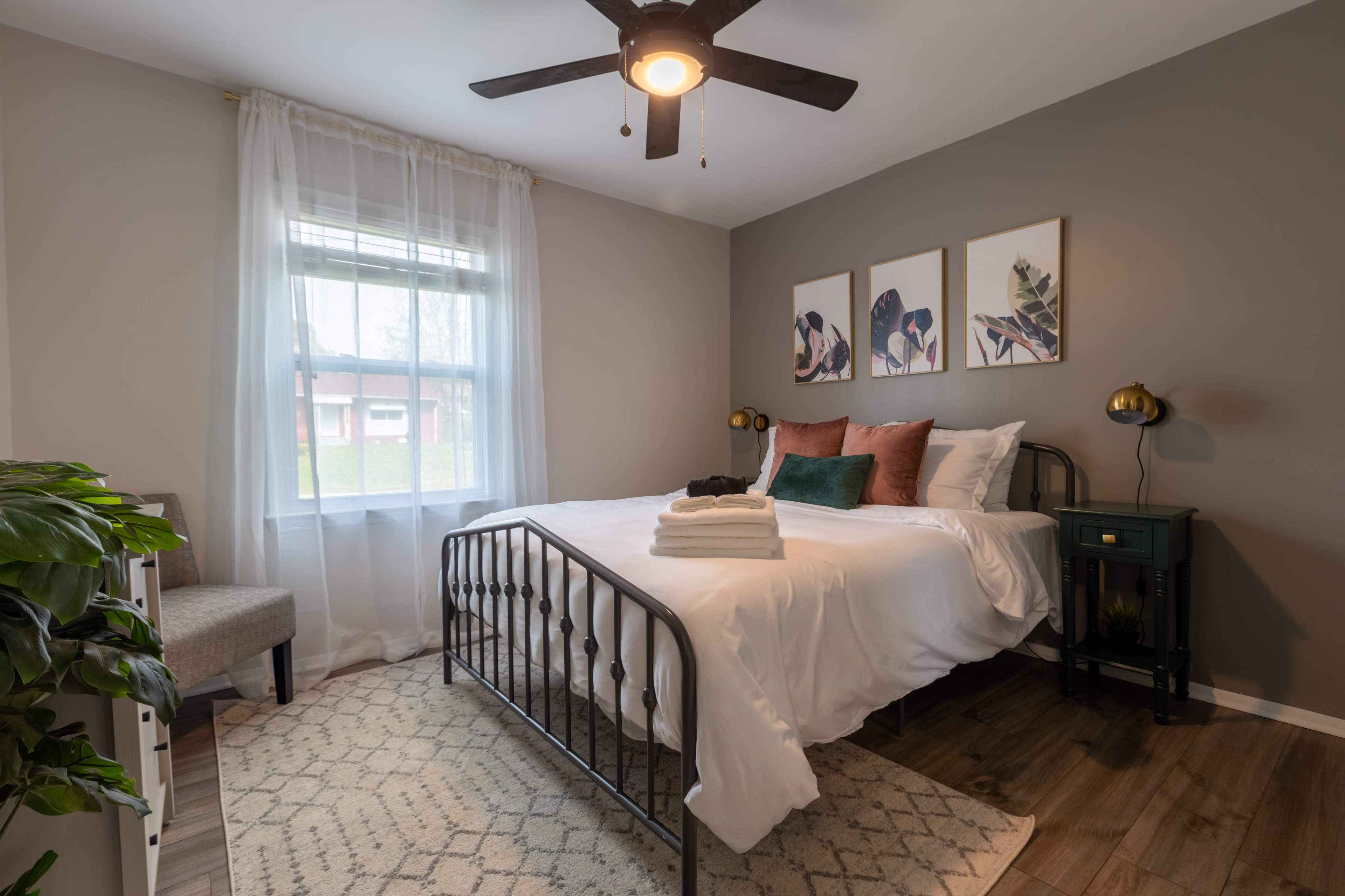 A neatly arranged bedroom featuring a bed with white bedding, a small green nightstand, and artwork on the walls, illuminated by natural light from a window.