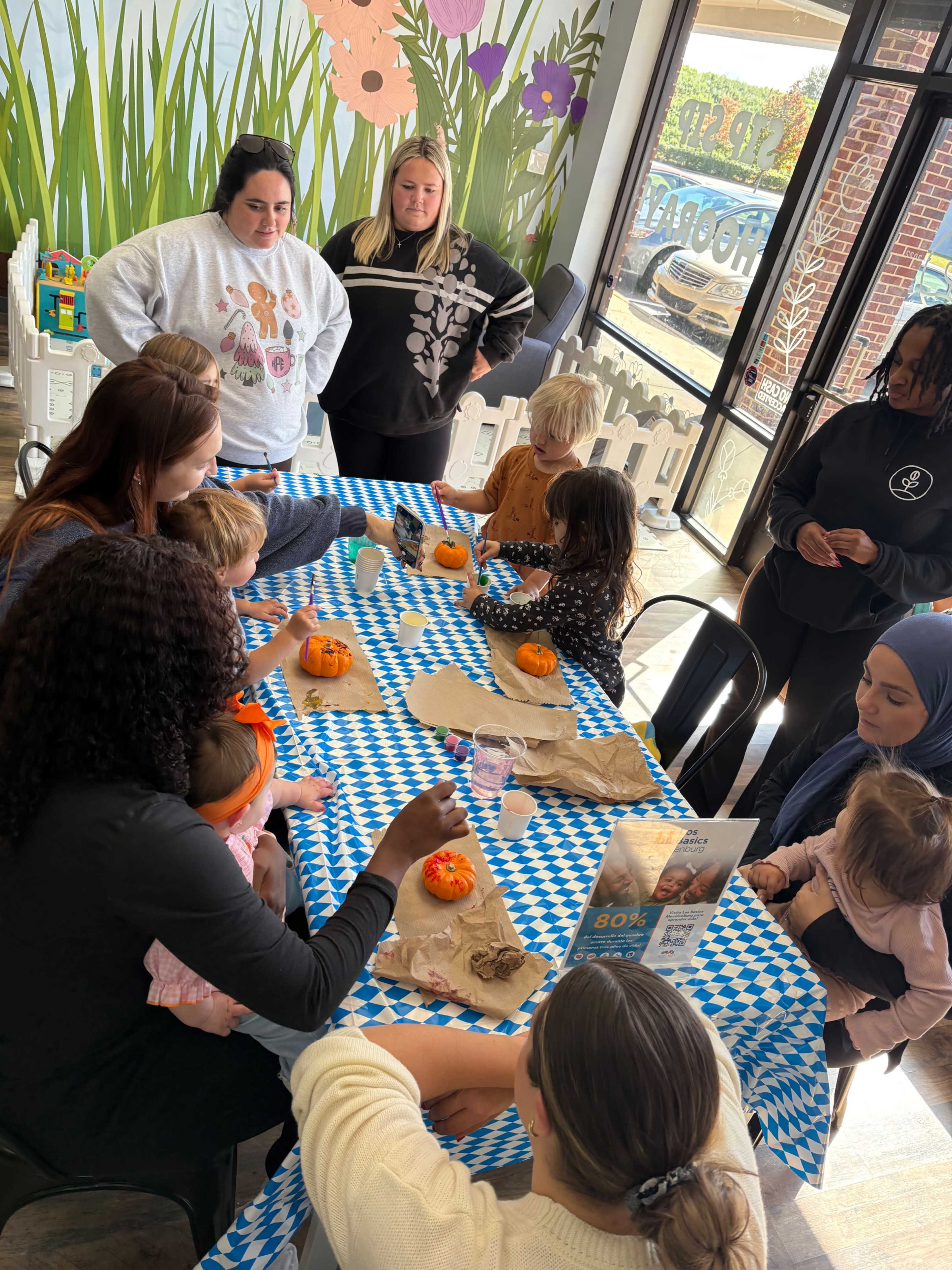 A group of children and adults are gathered around a blue-and-white tablecloth, engaged in pumpkin decorating activities in a brightly lit room with flower-themed decor.