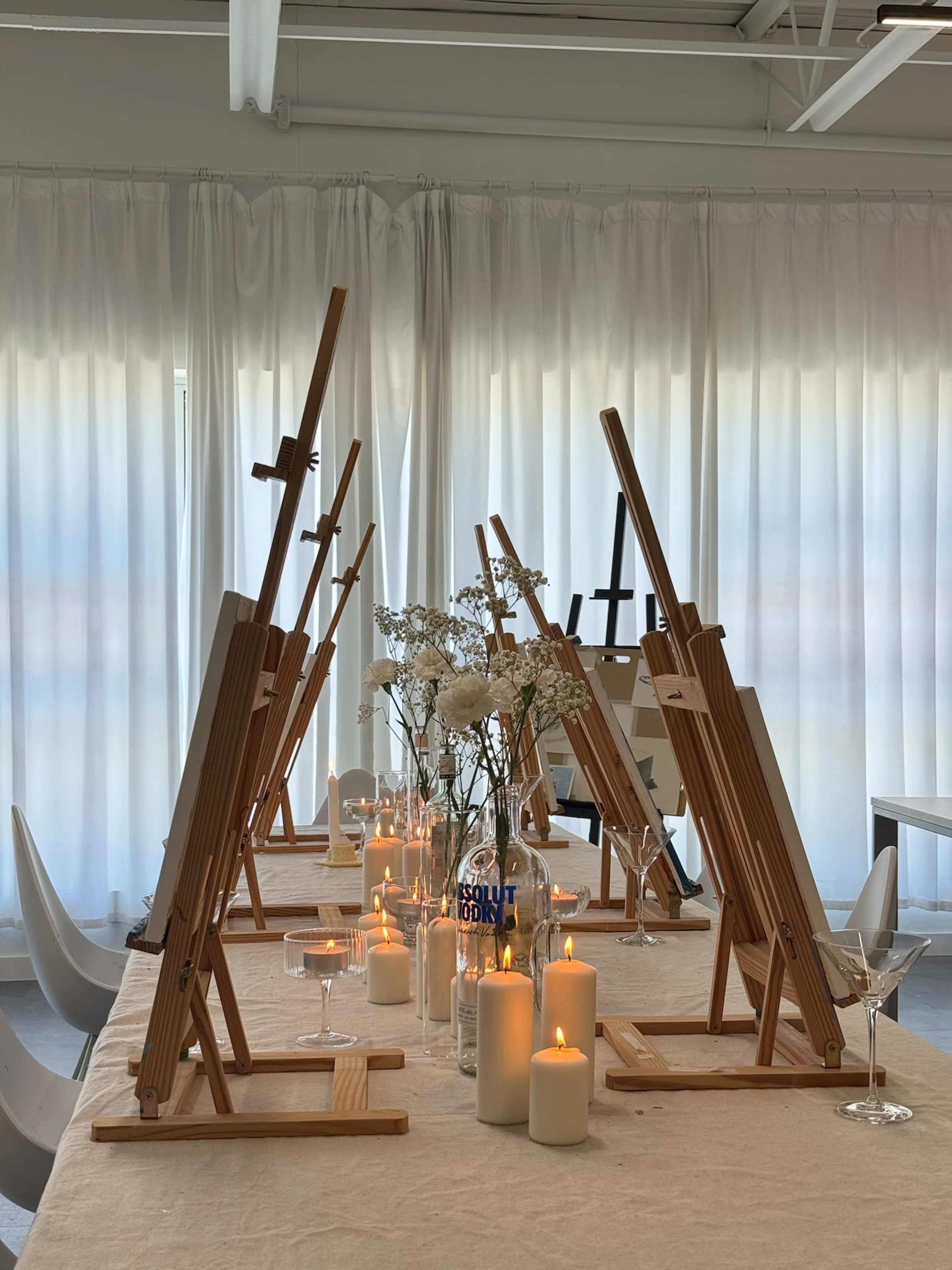 The image shows a setup of wooden easels arranged along a table, with candles and floral decorations in a well-lit room.