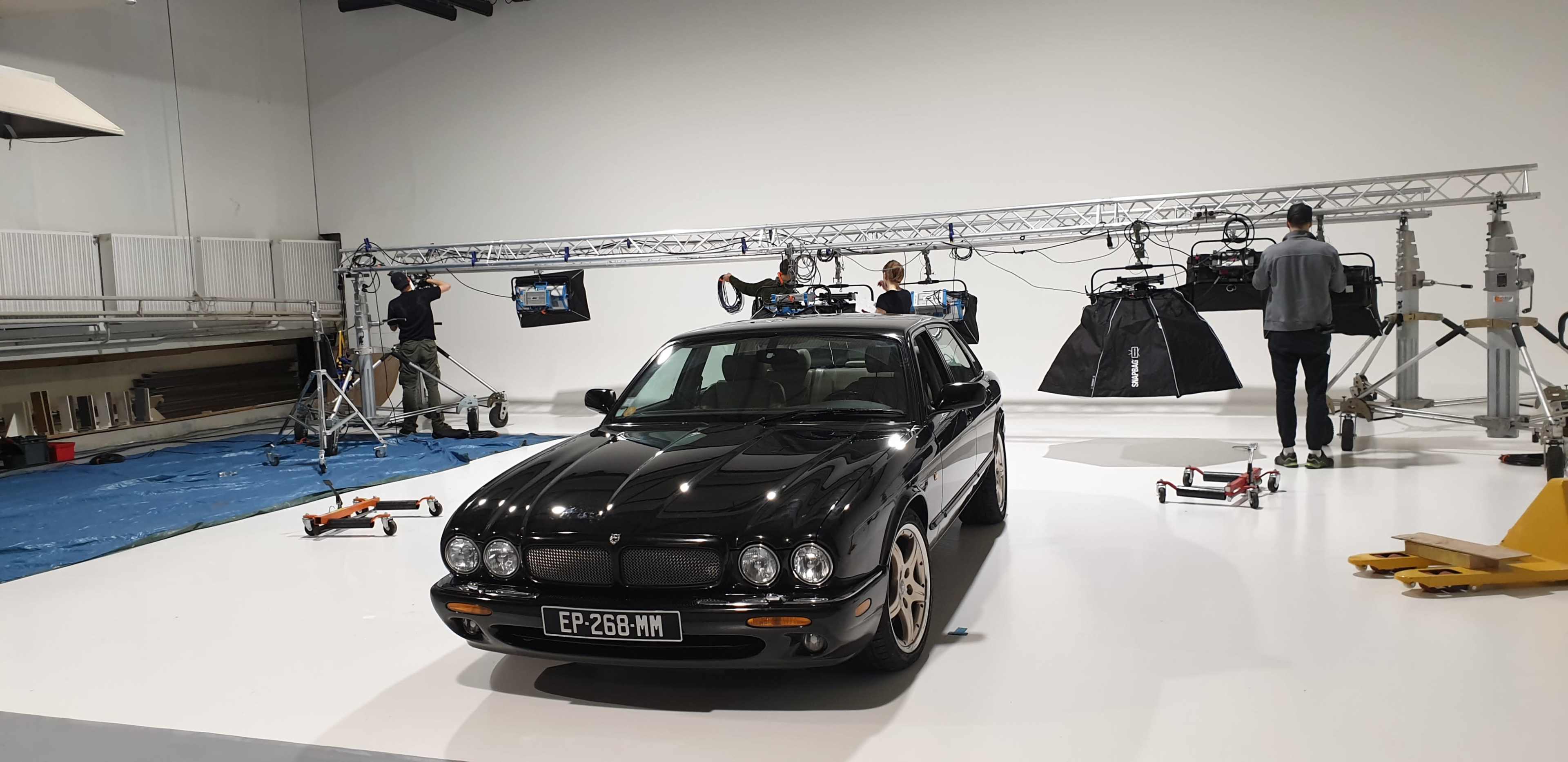 A black car is positioned in a studio, surrounded by lighting equipment and crew members setting up for a shoot.