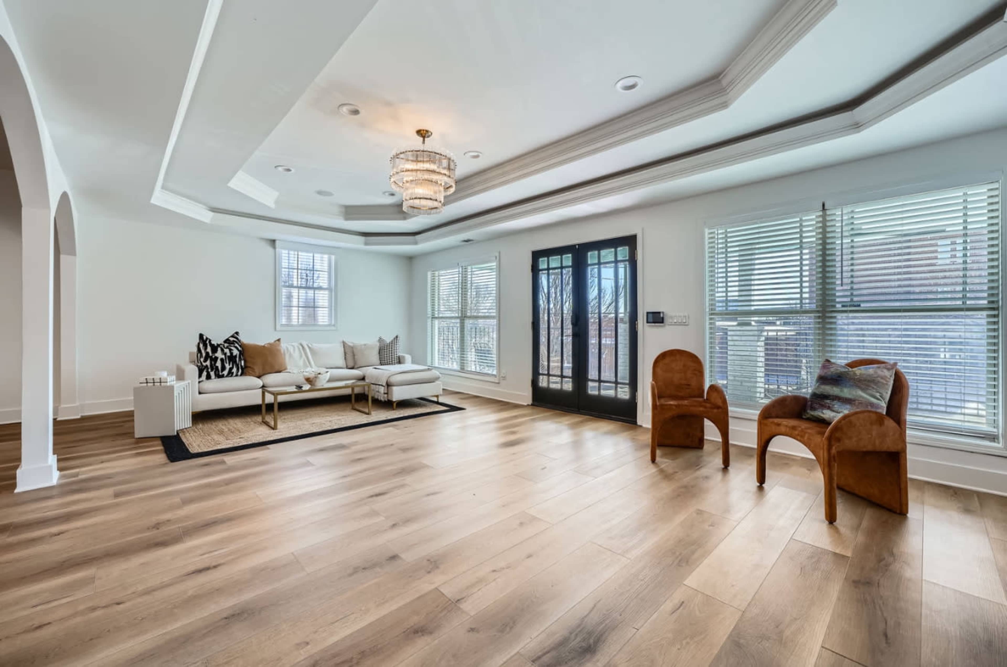 The image shows a spacious, well-lit living room featuring a large sofa, two chairs, a decorative area rug, and large windows with blinds.