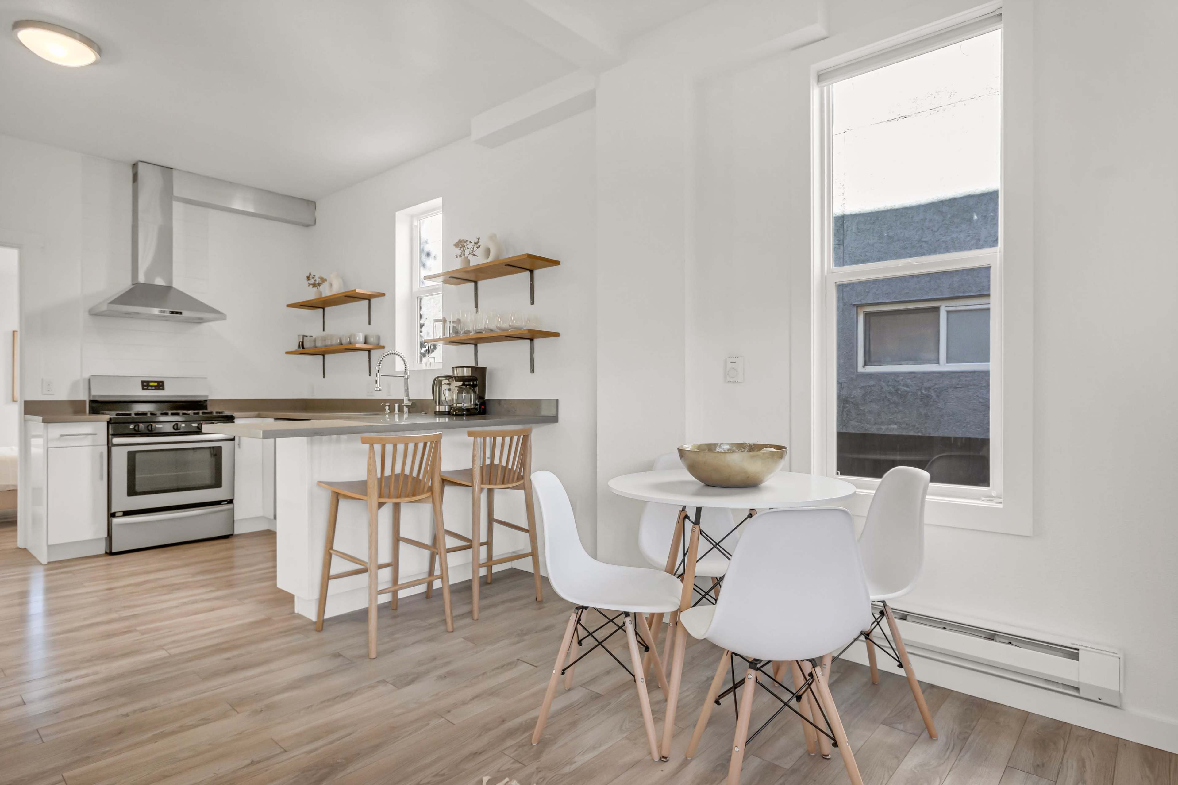 The image shows a modern kitchen and dining area featuring a stainless steel oven, wooden shelves, and a round dining table set with white chairs.