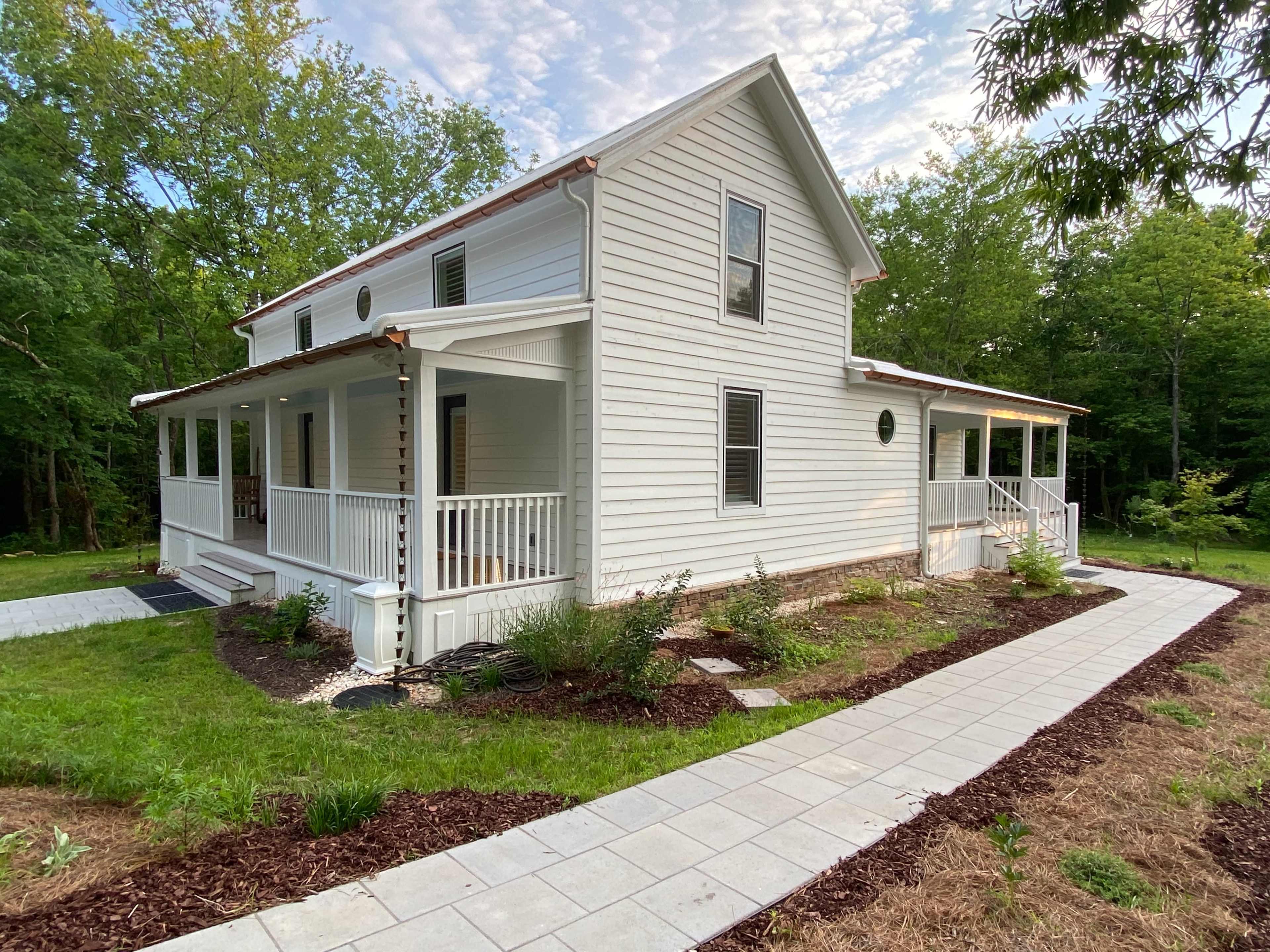 A two-story white house with a front porch, surrounded by green grass and small trees, with a pathway made of light-colored stones.