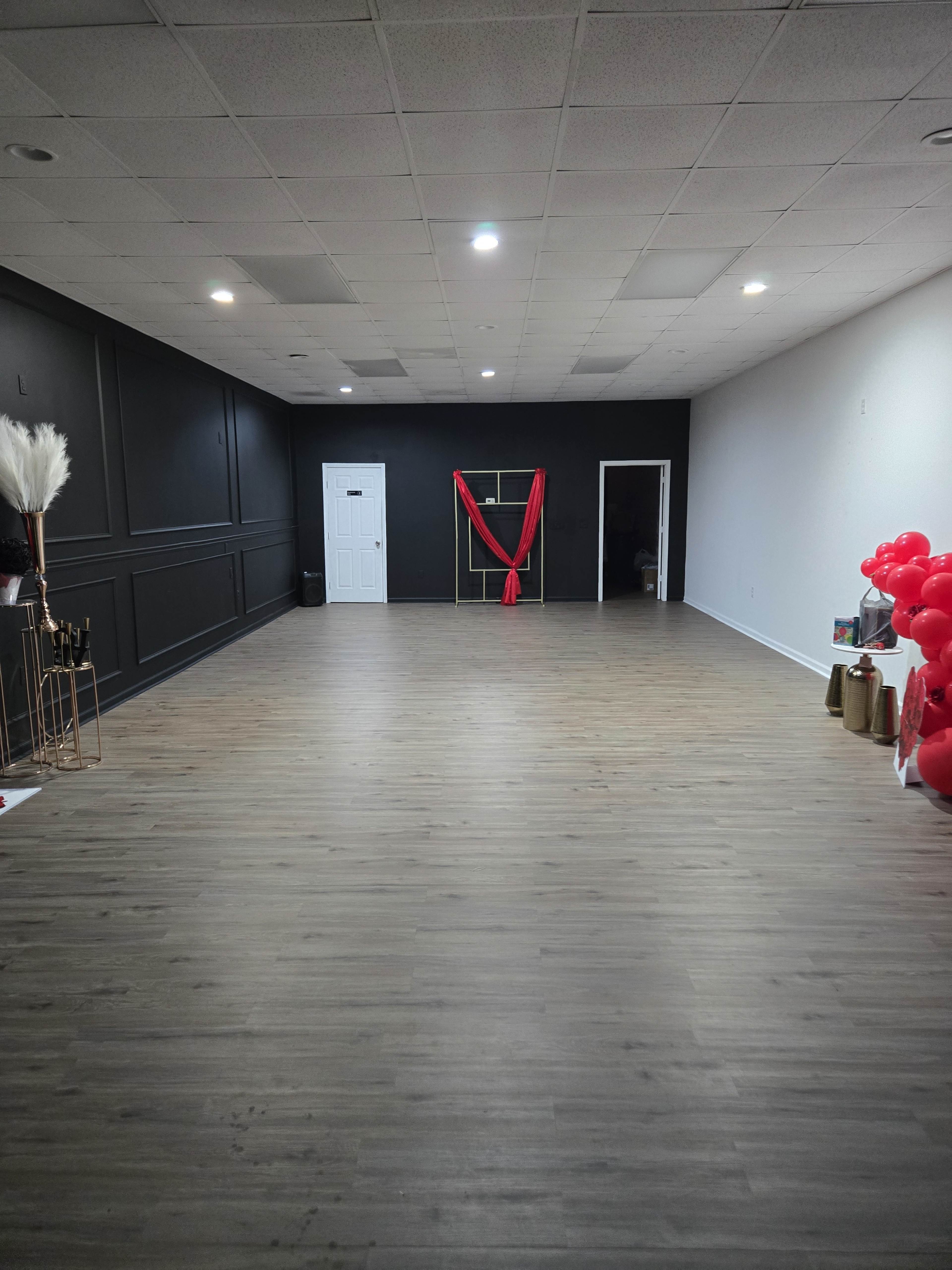 The image shows an empty room with wooden flooring, black and white walls, a red fabric draping in the center, and colorful decorations on one side.