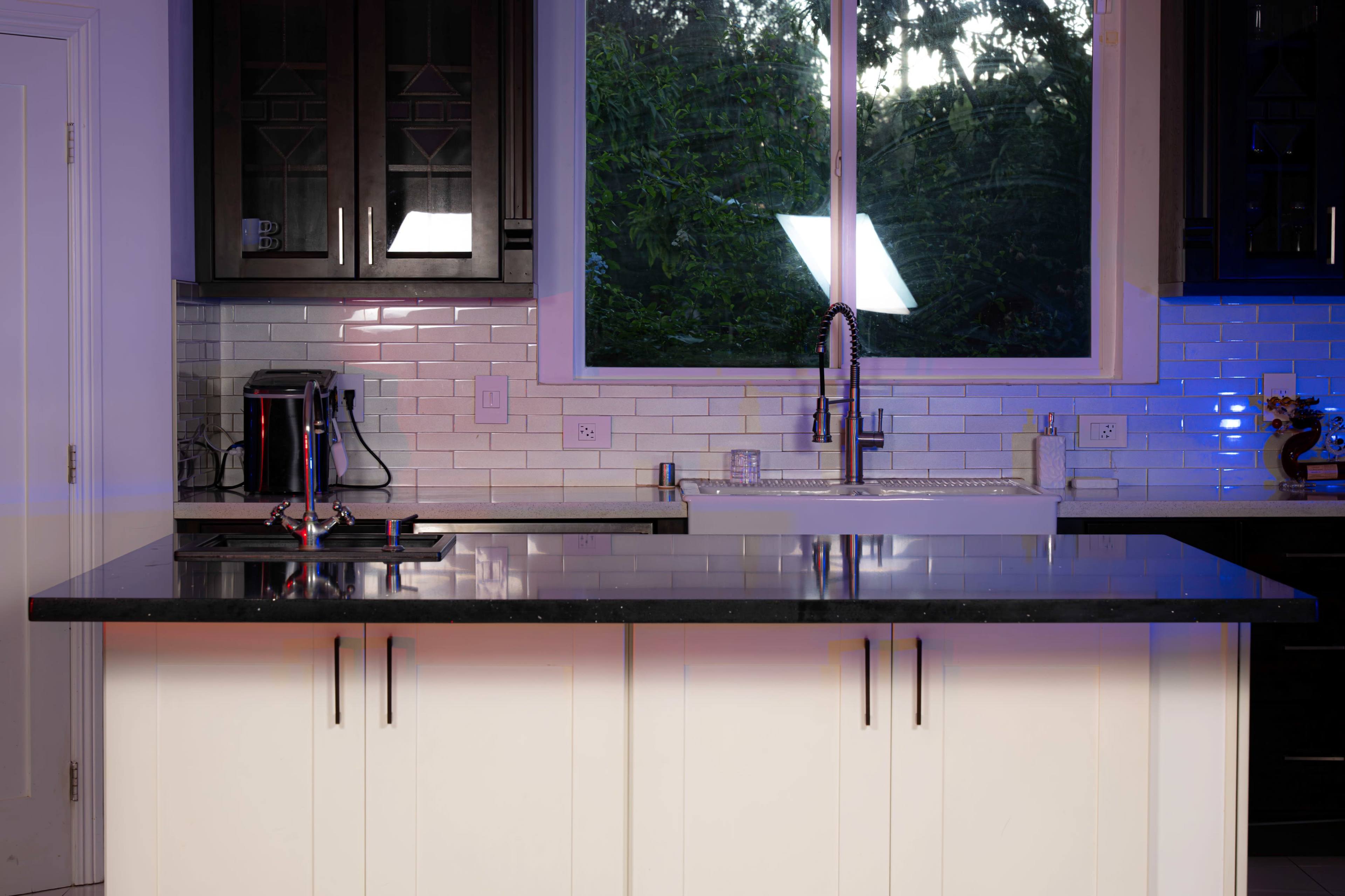 A modern kitchen features a dark countertop and cabinetry, illuminated by natural light from a window and accent lighting.