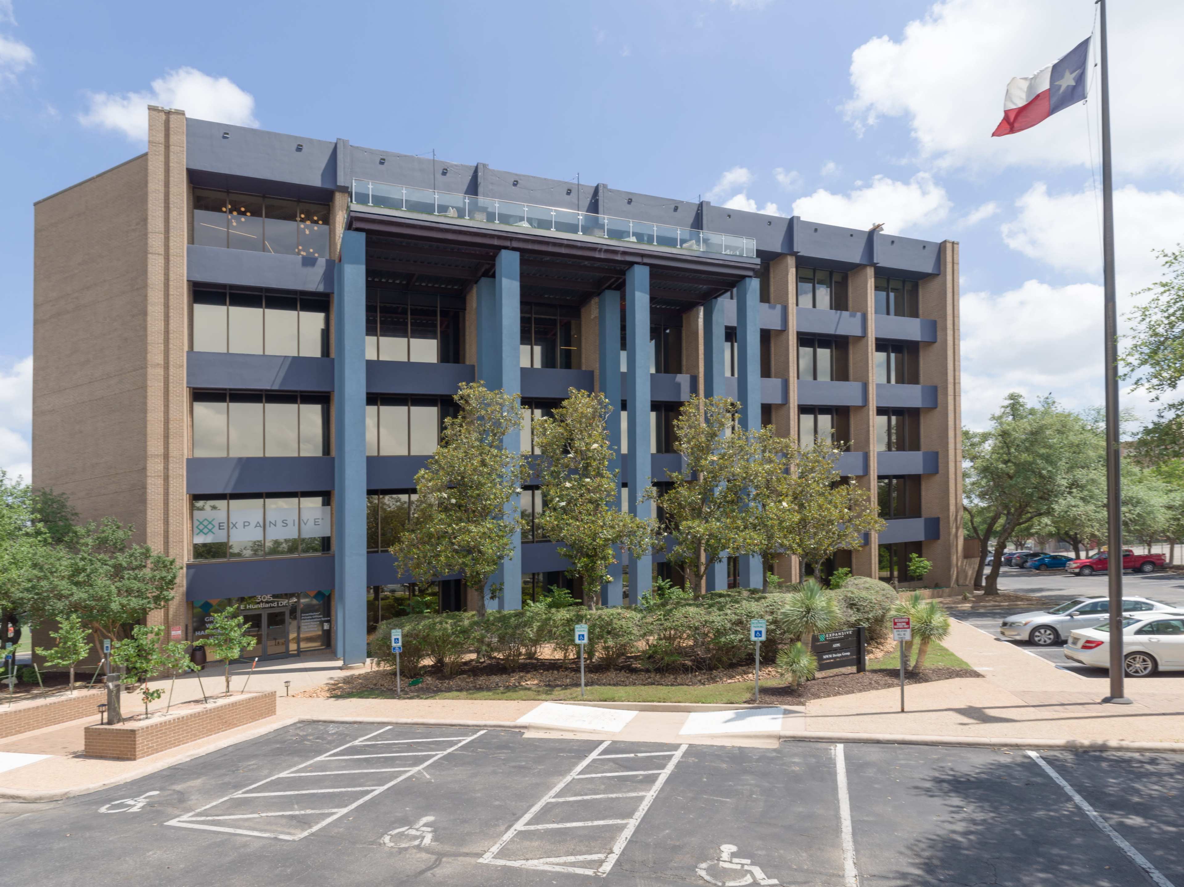 The image shows a four-story office building with dark blue and beige accents, surrounded by trees and parked cars, with a Texas flag on a pole nearby.