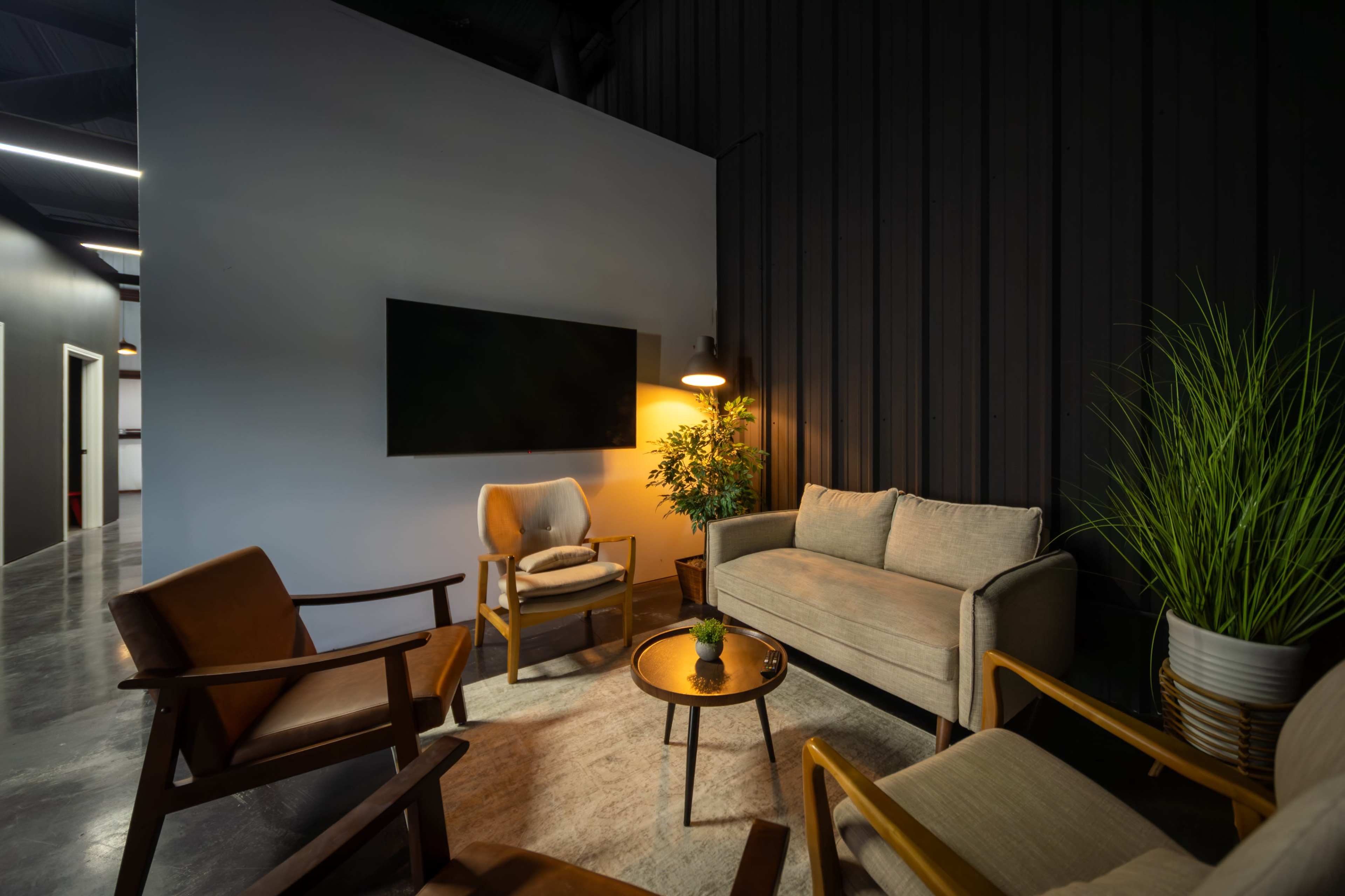 The image shows a modern lounge area featuring two armchairs, a couch, a round coffee table, a floor lamp, and a potted plant against a wall with a television.
