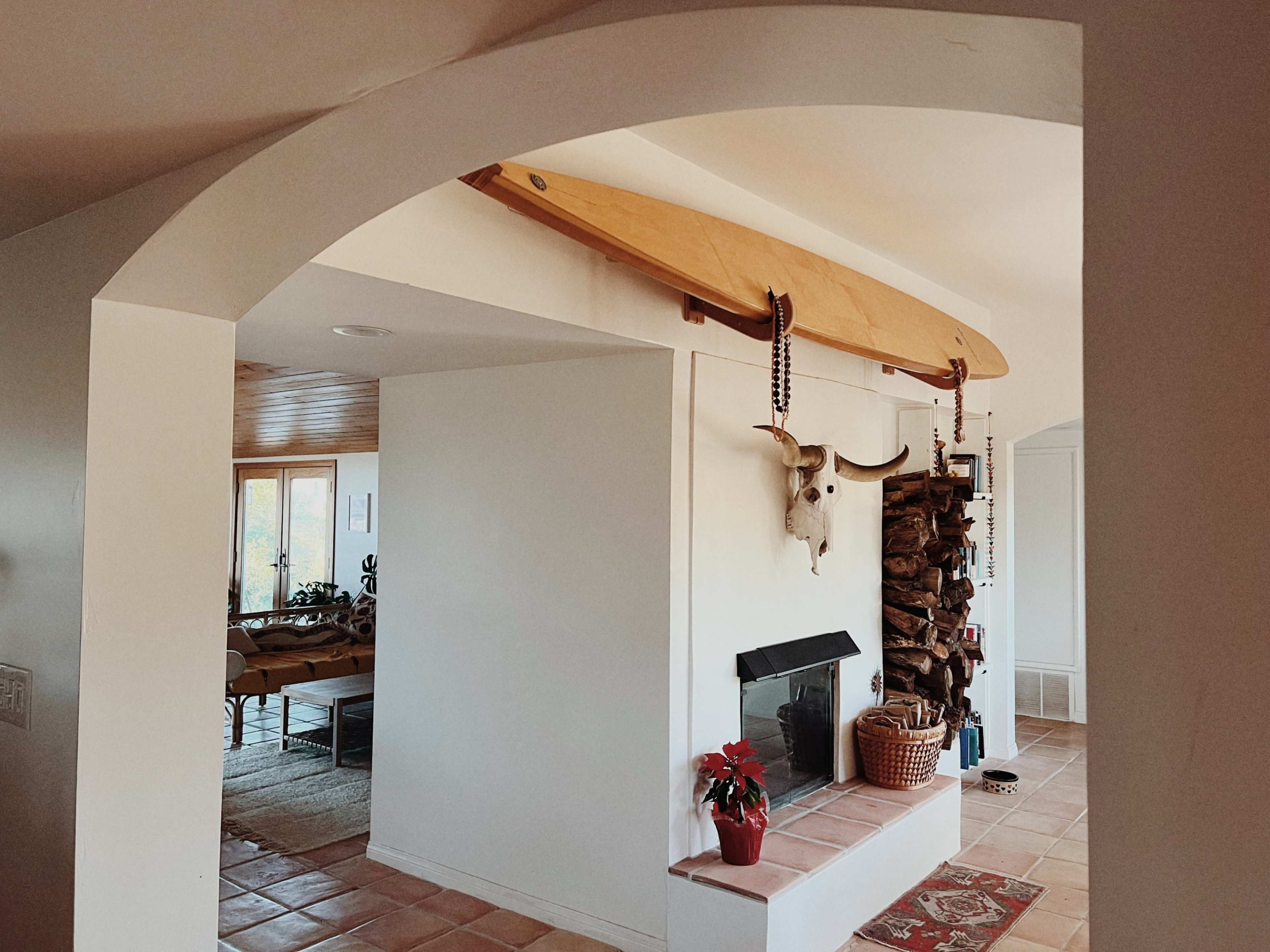 The image shows an interior space featuring a wooden surfboard mounted above a fireplace, flanked by a decorative cow skull and a basket of logs.