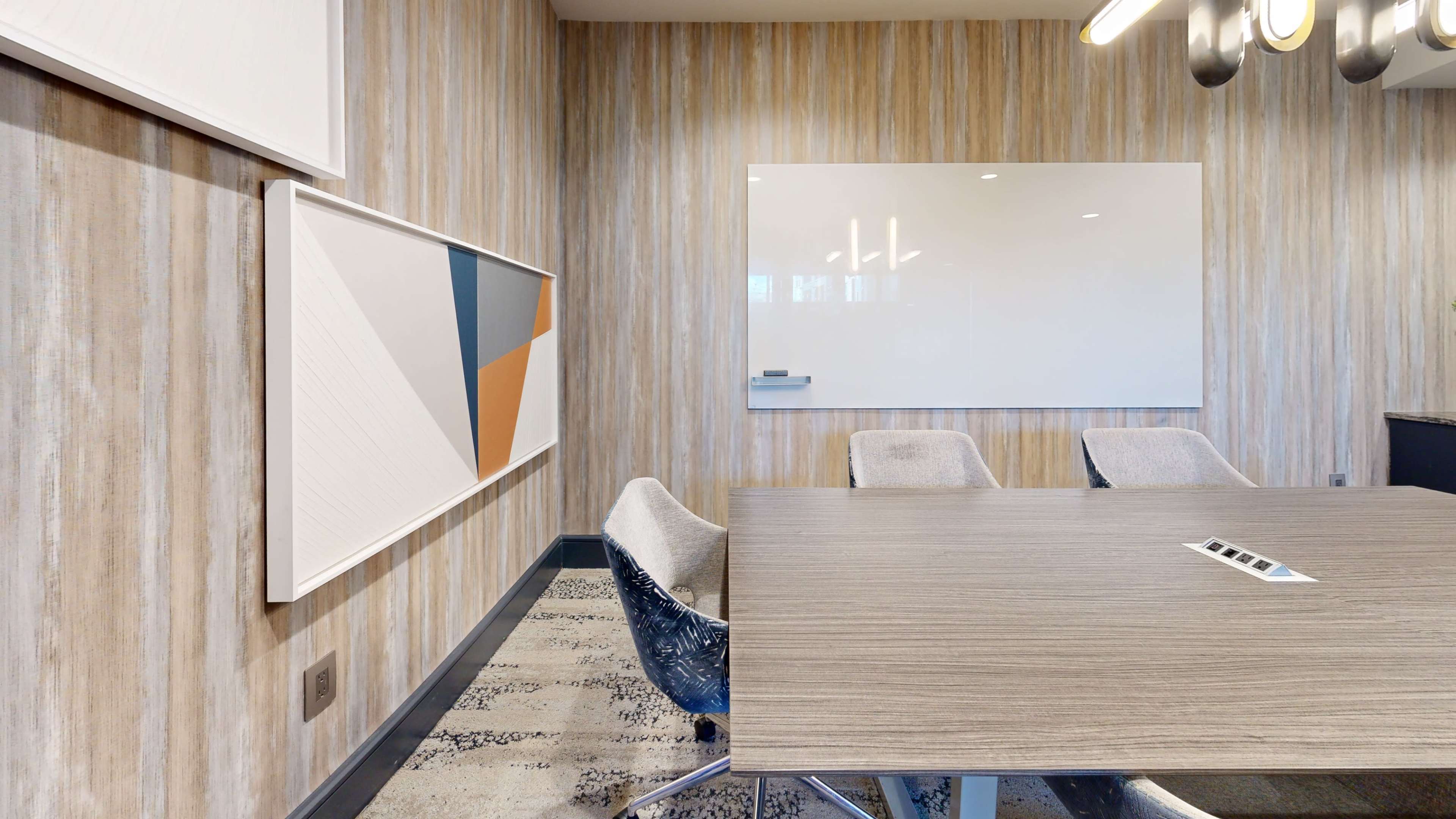 A modern meeting room features a wooden table surrounded by gray chairs, with a large whiteboard and framed artwork on the wall.