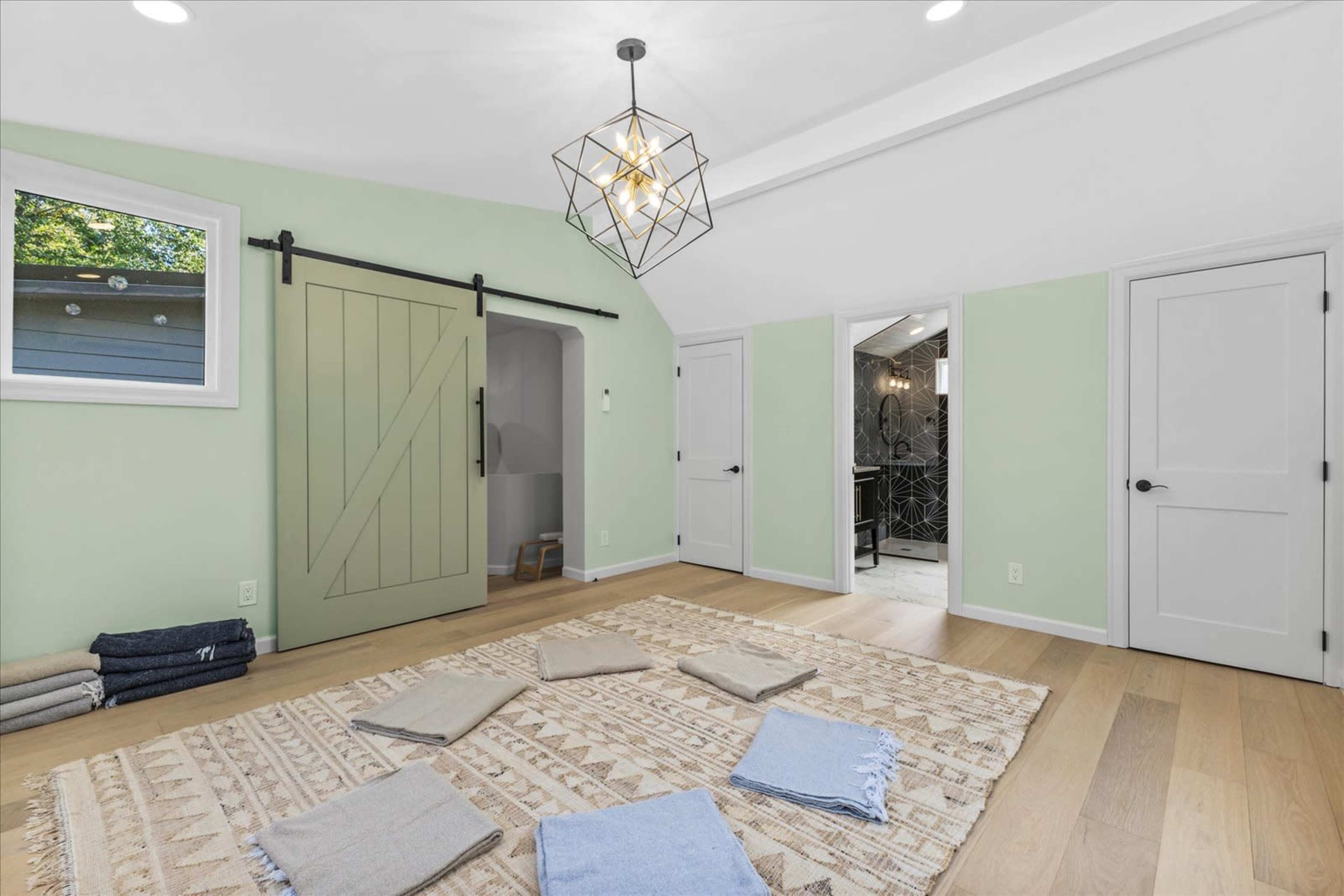 The image shows a light green room with a patterned rug on the wooden floor, surrounded by folded blankets and featuring a sliding barn door and two white doors.