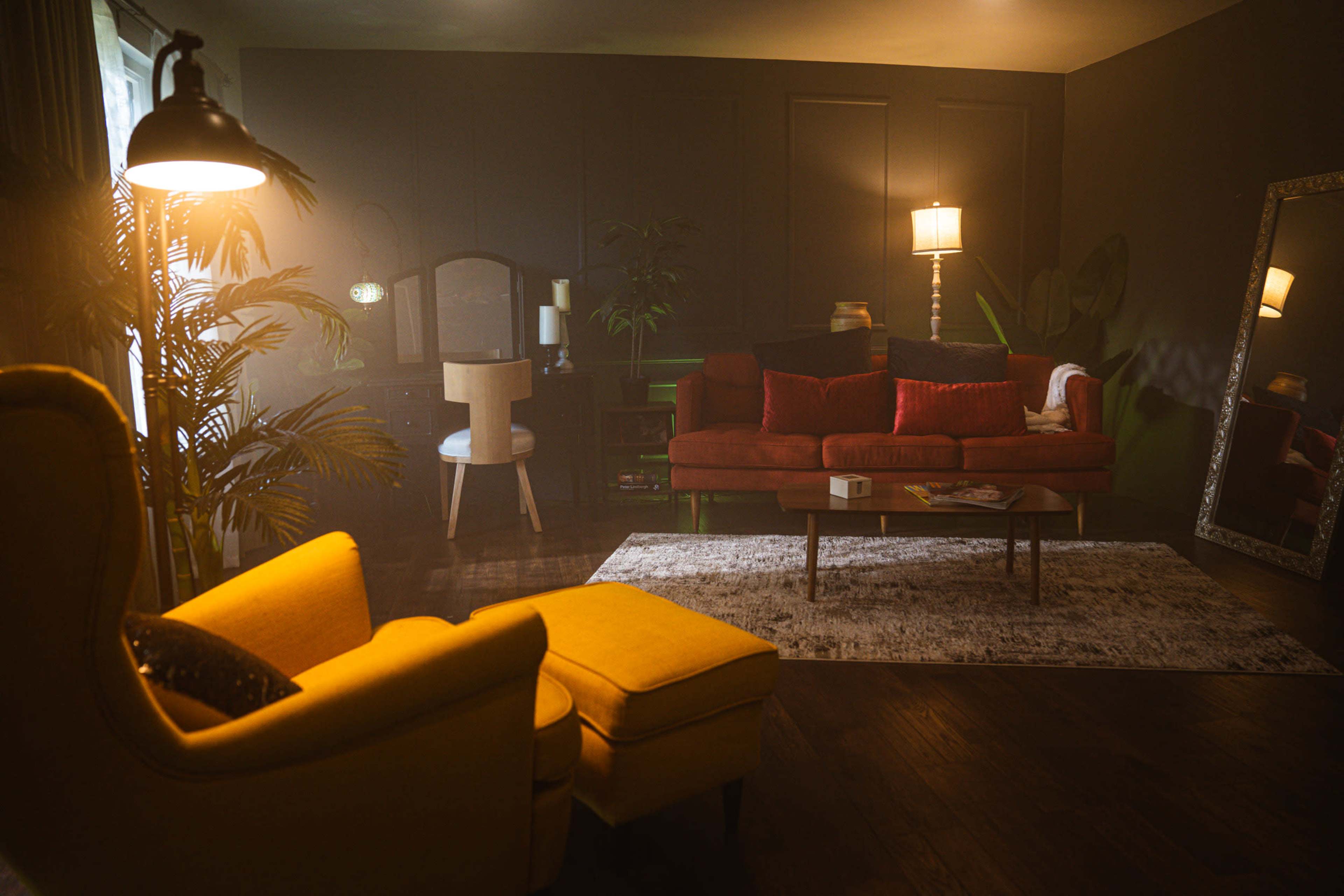 A living room features a yellow armchair and ottoman, a dark sofa with red pillows, a large mirror, and plants surrounding a coffee table on a rug.